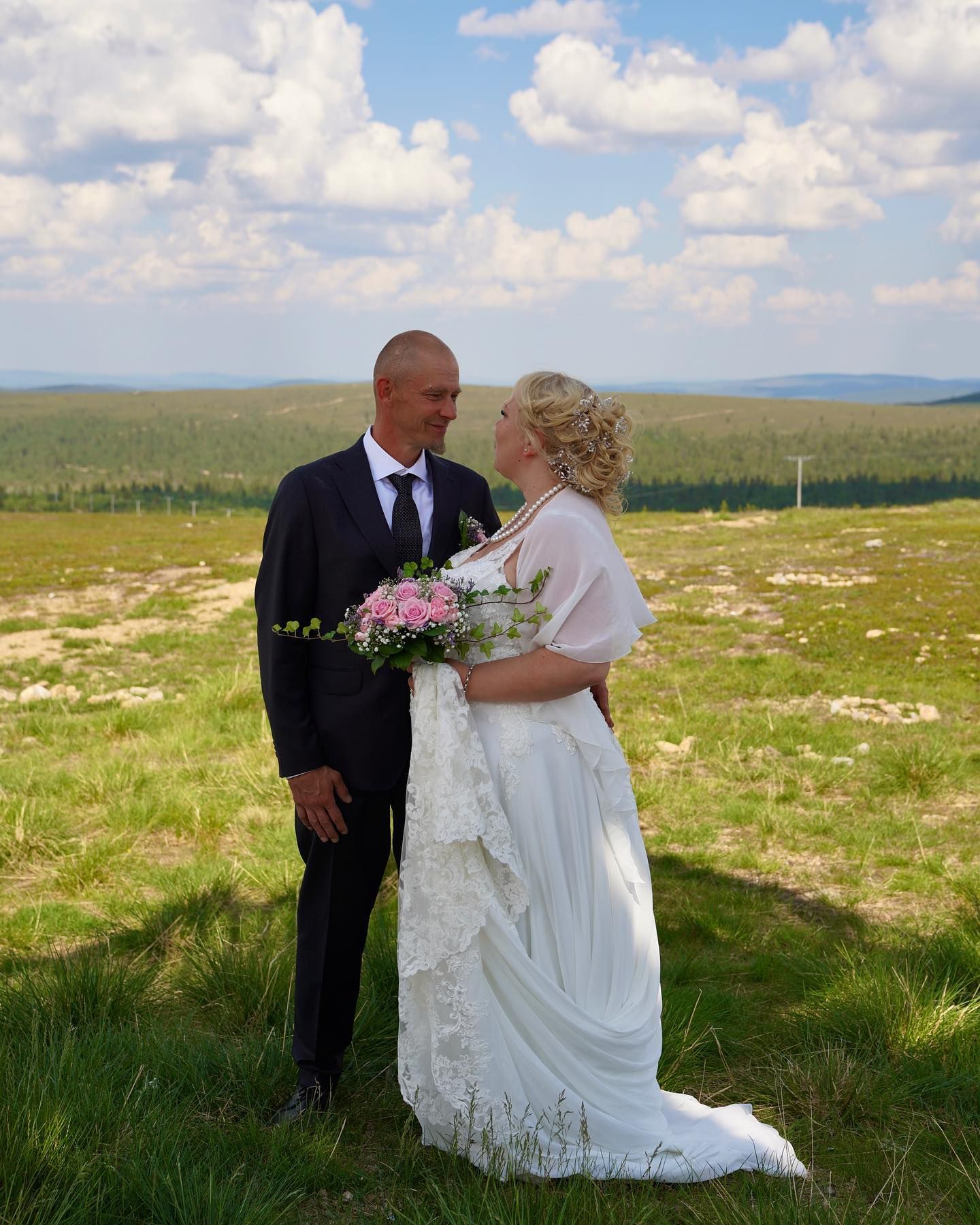 A couple in wedding attire embraces on a hilltop meadow with rolling hills and puffy clouds in the background.