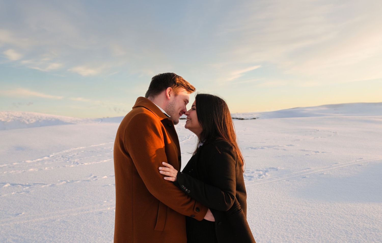 Couple embracing in snowy landscape during sunset with mountains in background.