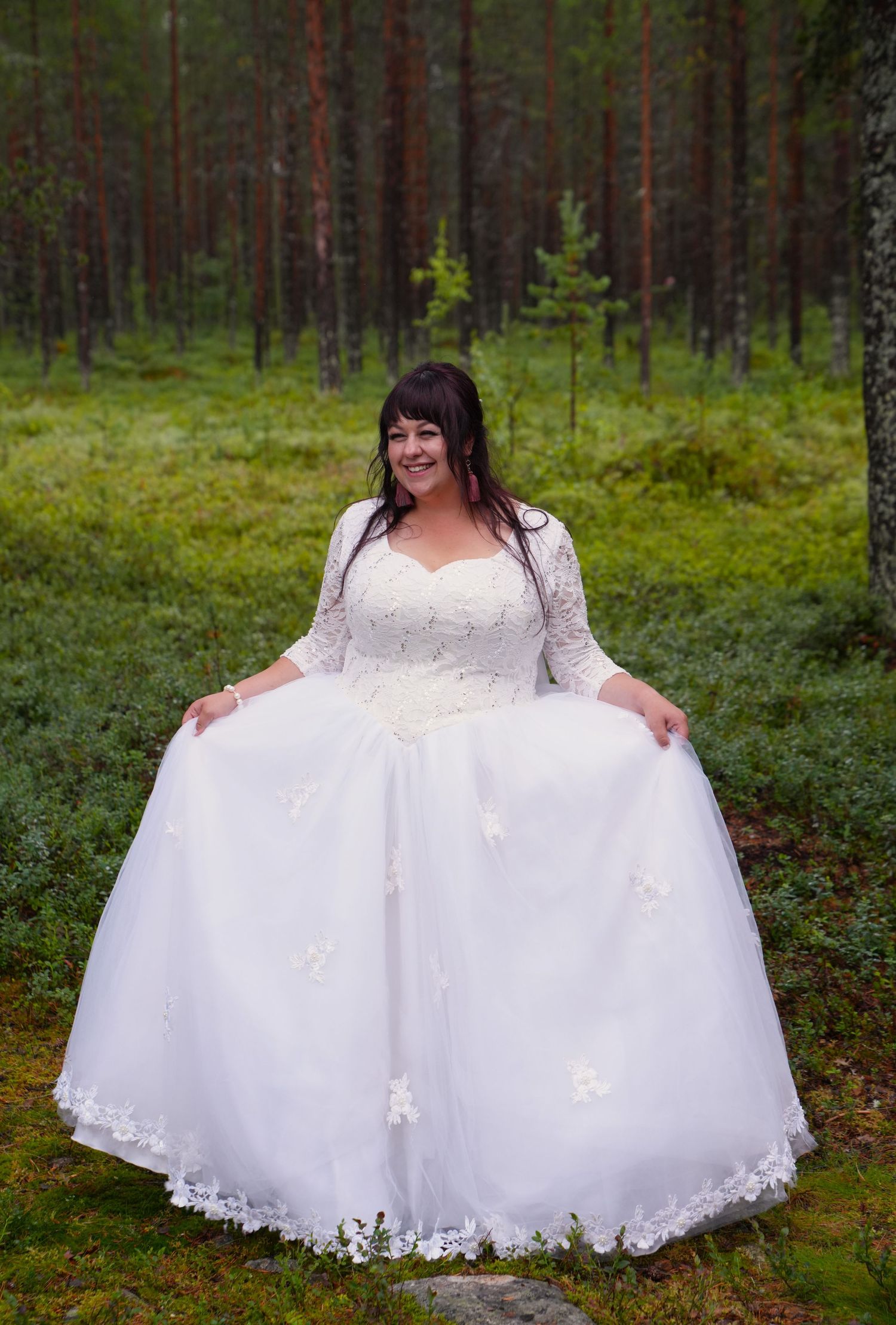 Person in white lace wedding gown standing in lush green forest setting.