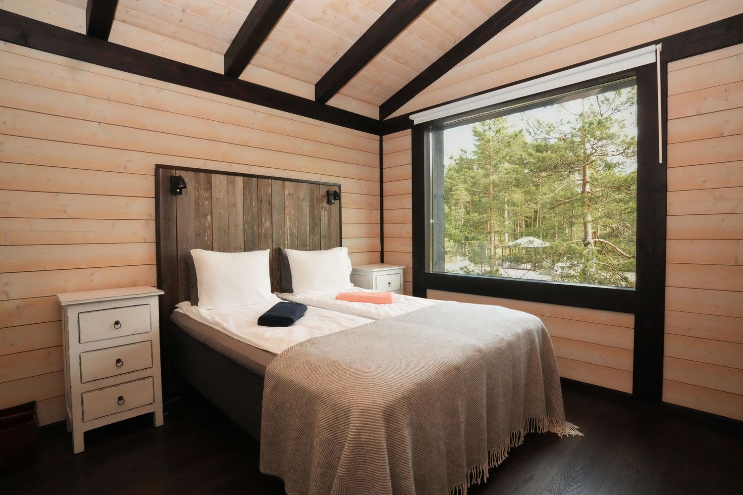 Modern bedroom in wooden cabin with large window overlooking forest, featuring rustic decor and white bedding.