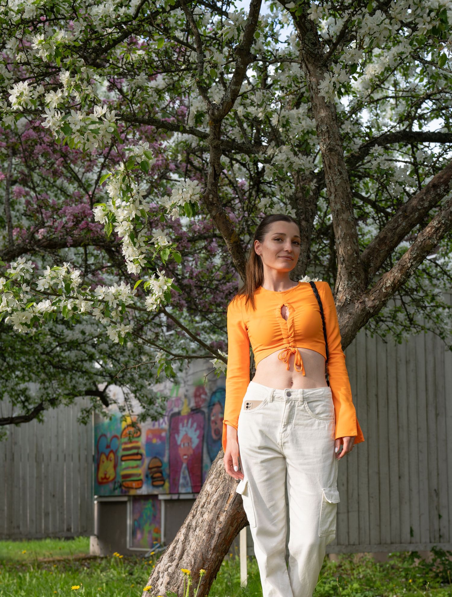 Woman in bright orange crop top and white pants posing near blooming spring tree.