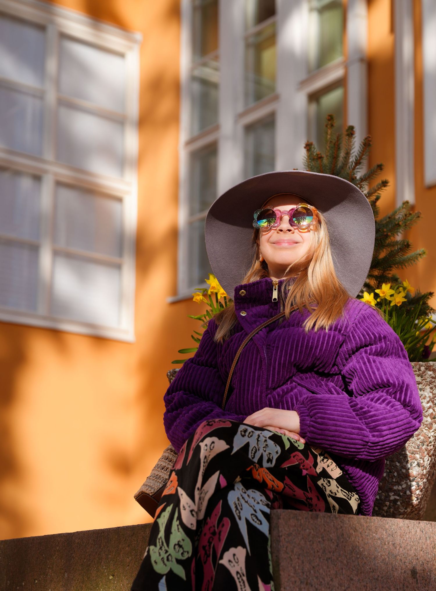 Stylish person in purple sweater and wide-brimmed hat sitting against orange building exterior.