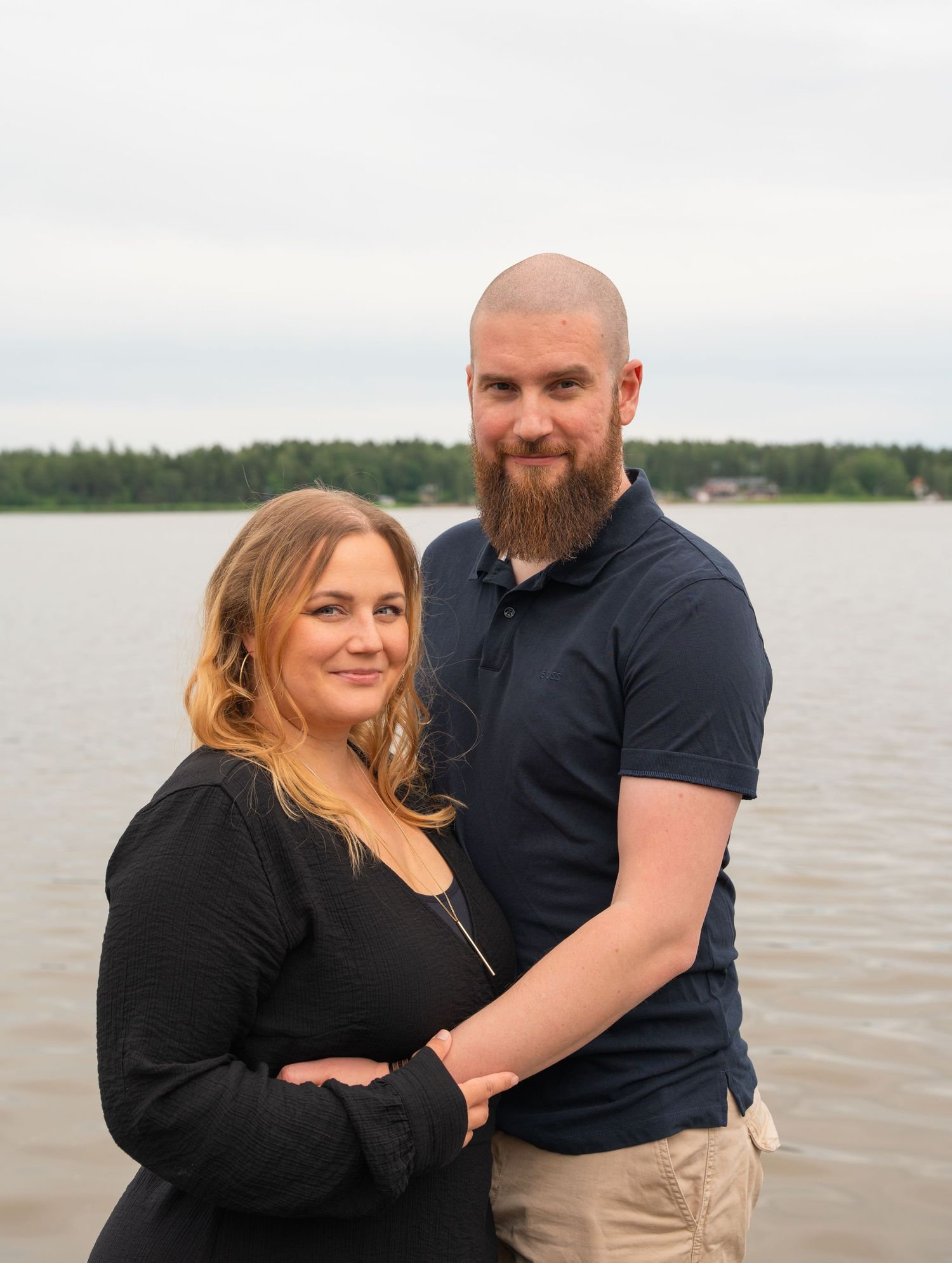 A couple embraces while posing together by a lake on an overcast day, with trees visible in the distance.