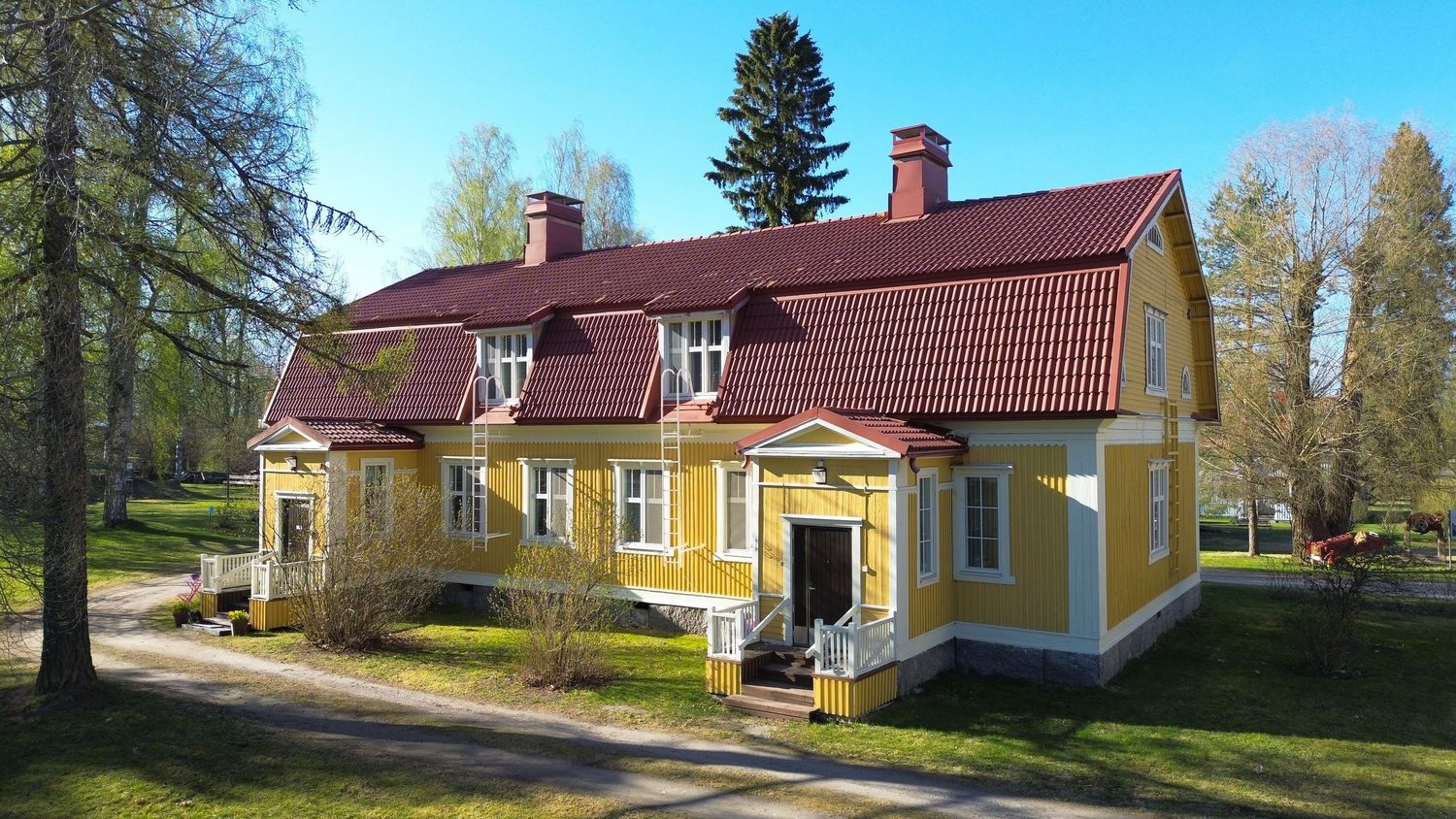 Yellow Finnish wooden manor house with red roof surrounded by tall trees on a sunny spring day.