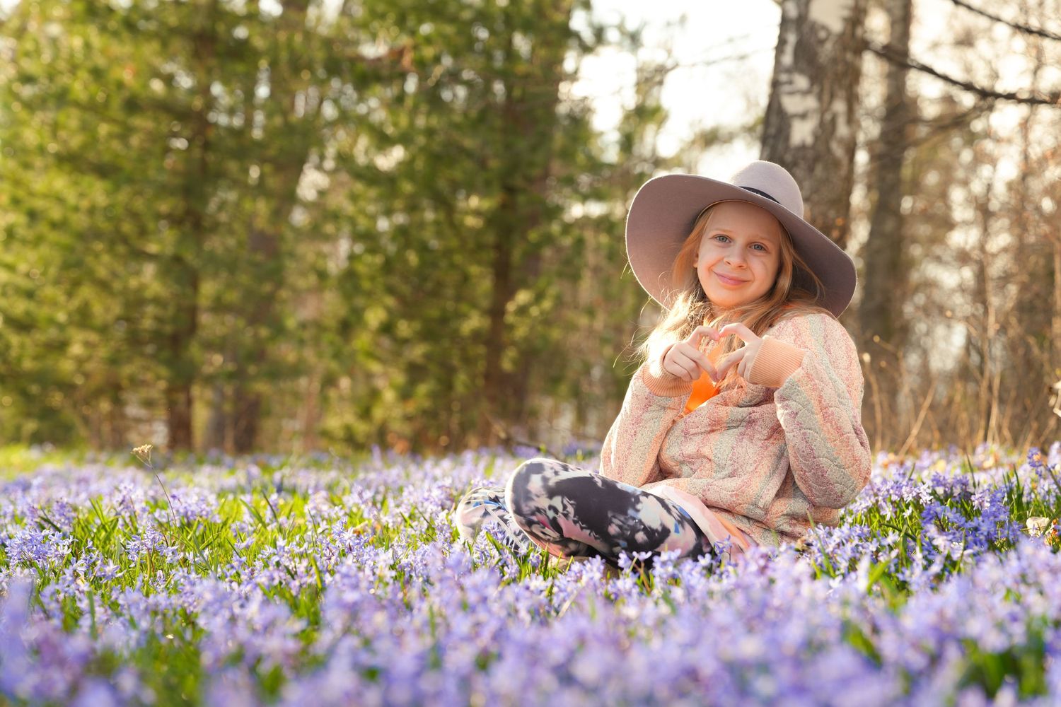 Child in hat sitting among purple wildflowers in a spring forest setting.