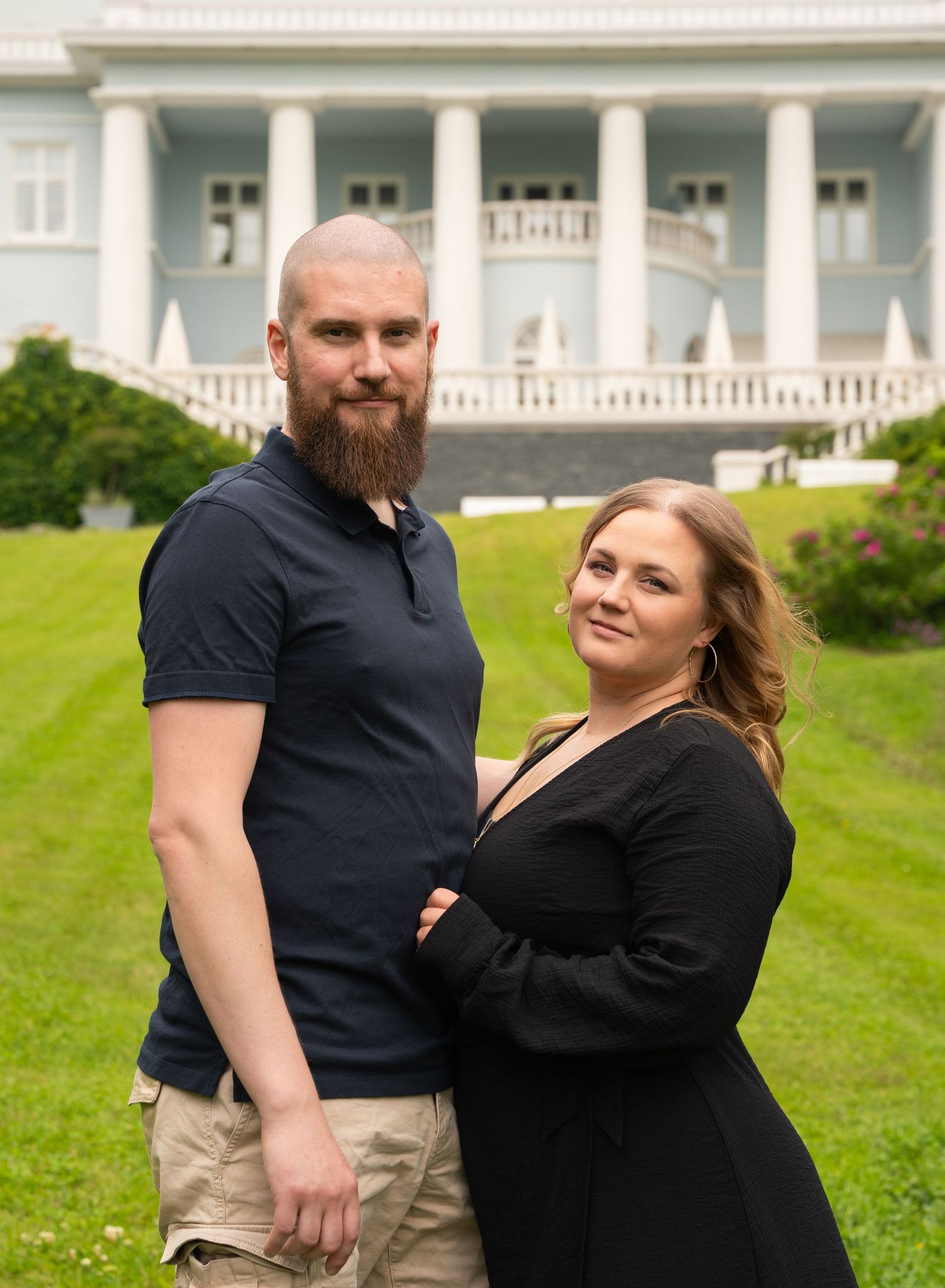 A couple embraces and laughs together on a manicured lawn with a grand white building in the background.