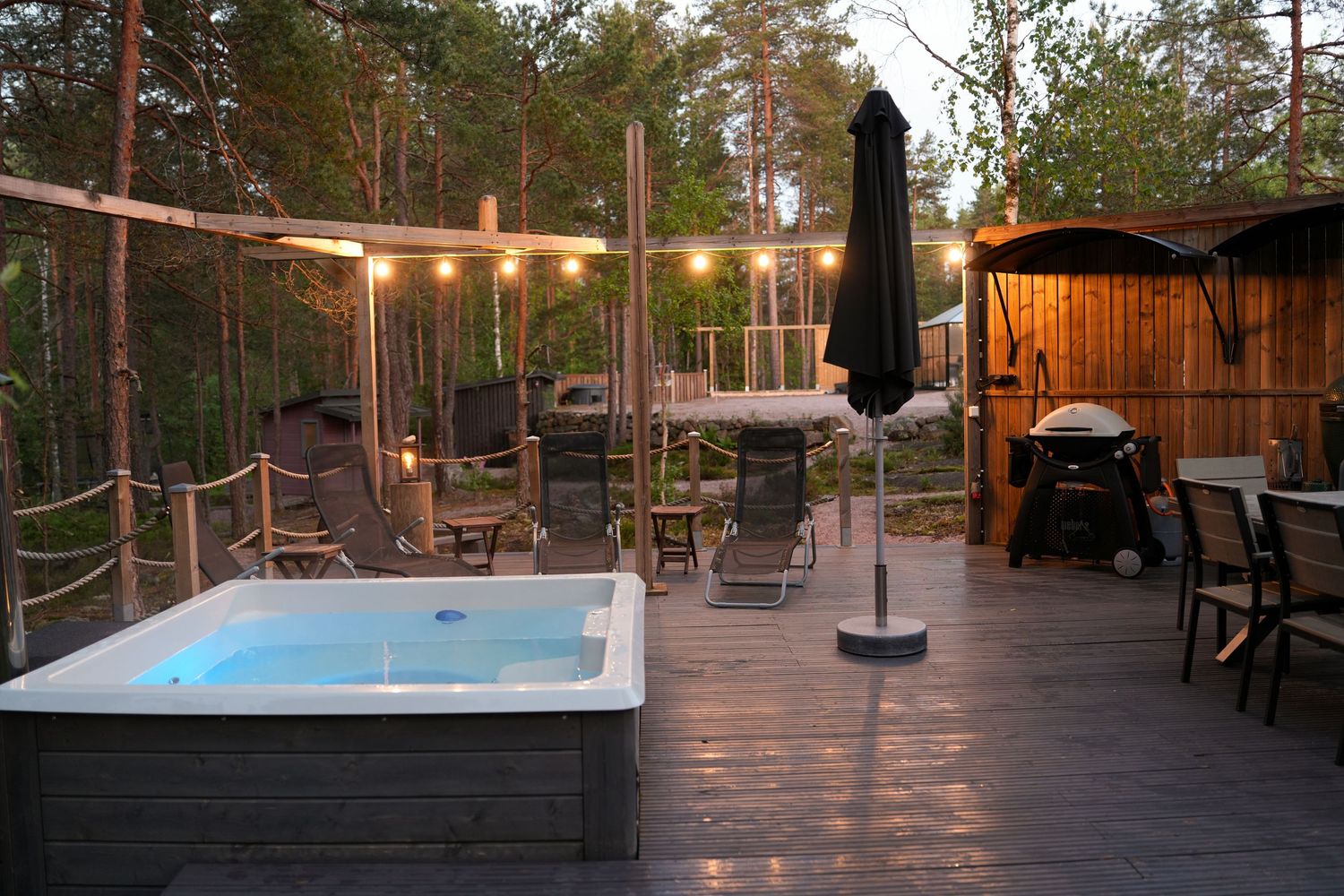Outdoor deck with hot tub and string lights features wooden railings and seating areas in a serene forest setting at dusk.