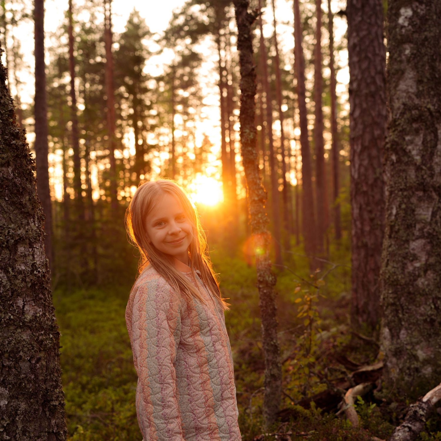 Person in a floral dress stands among tall pine trees at golden sunset in a forest landscape.