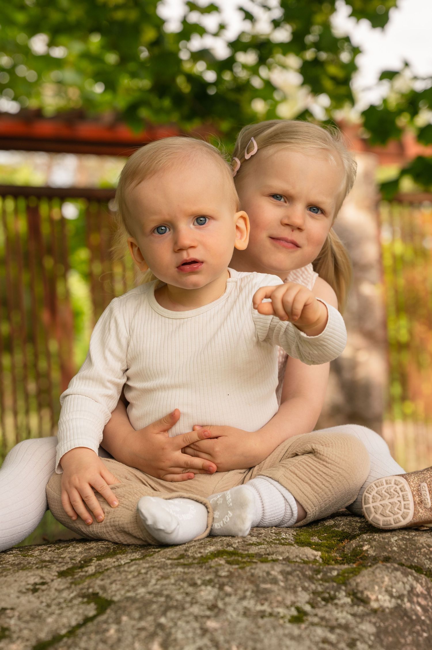 Two toddlers in white and beige outfits sit together on a stone surface in a garden setting.