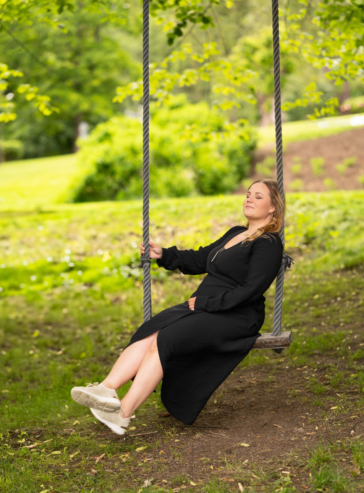 Someone wearing a black dress swings joyfully on a rope swing in a lush green outdoor setting during spring or summer.
