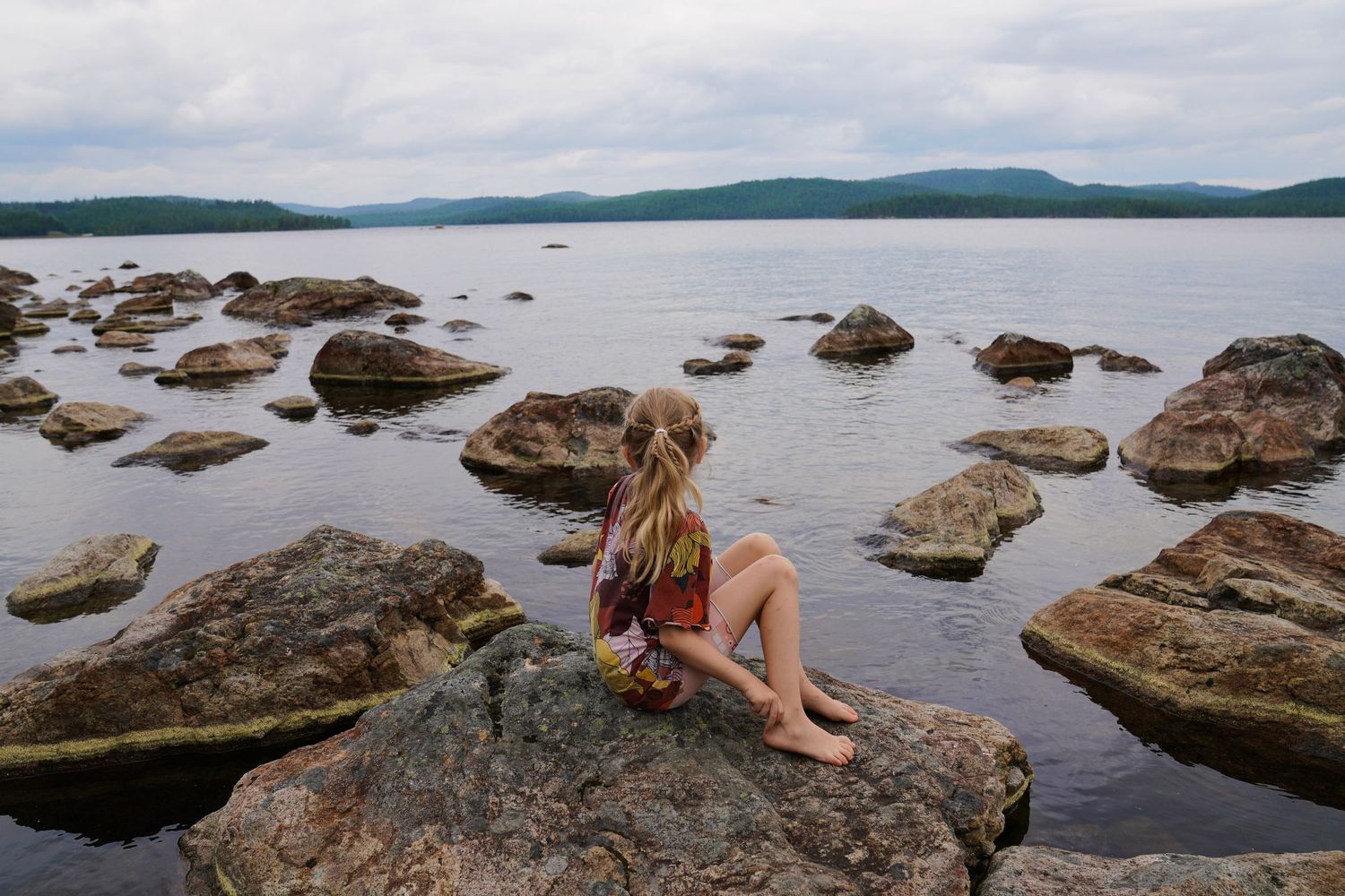 Woman relaxing on rocky shoreline overlooking calm lake and distant mountains.