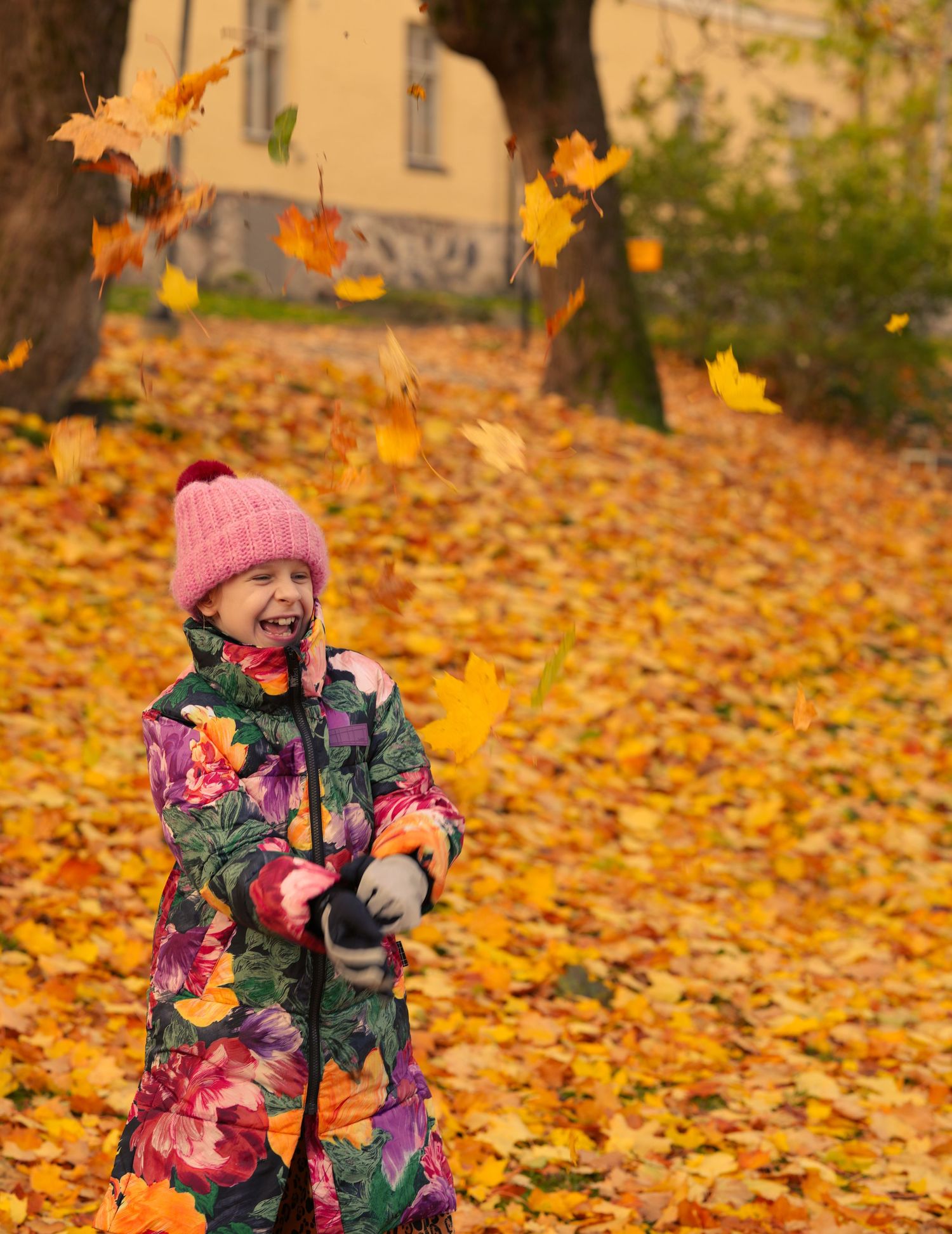 Child in floral coat and pink hat playing with falling autumn leaves.
