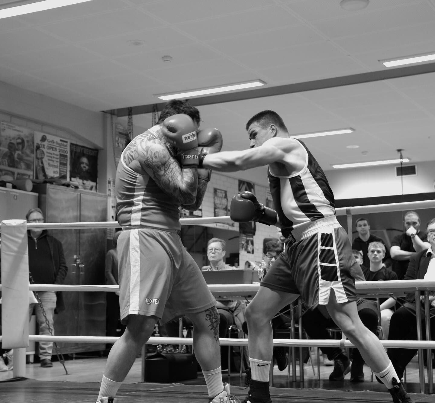 Two boxers spar in a boxing ring at a gym while spectators watch from outside the ropes.