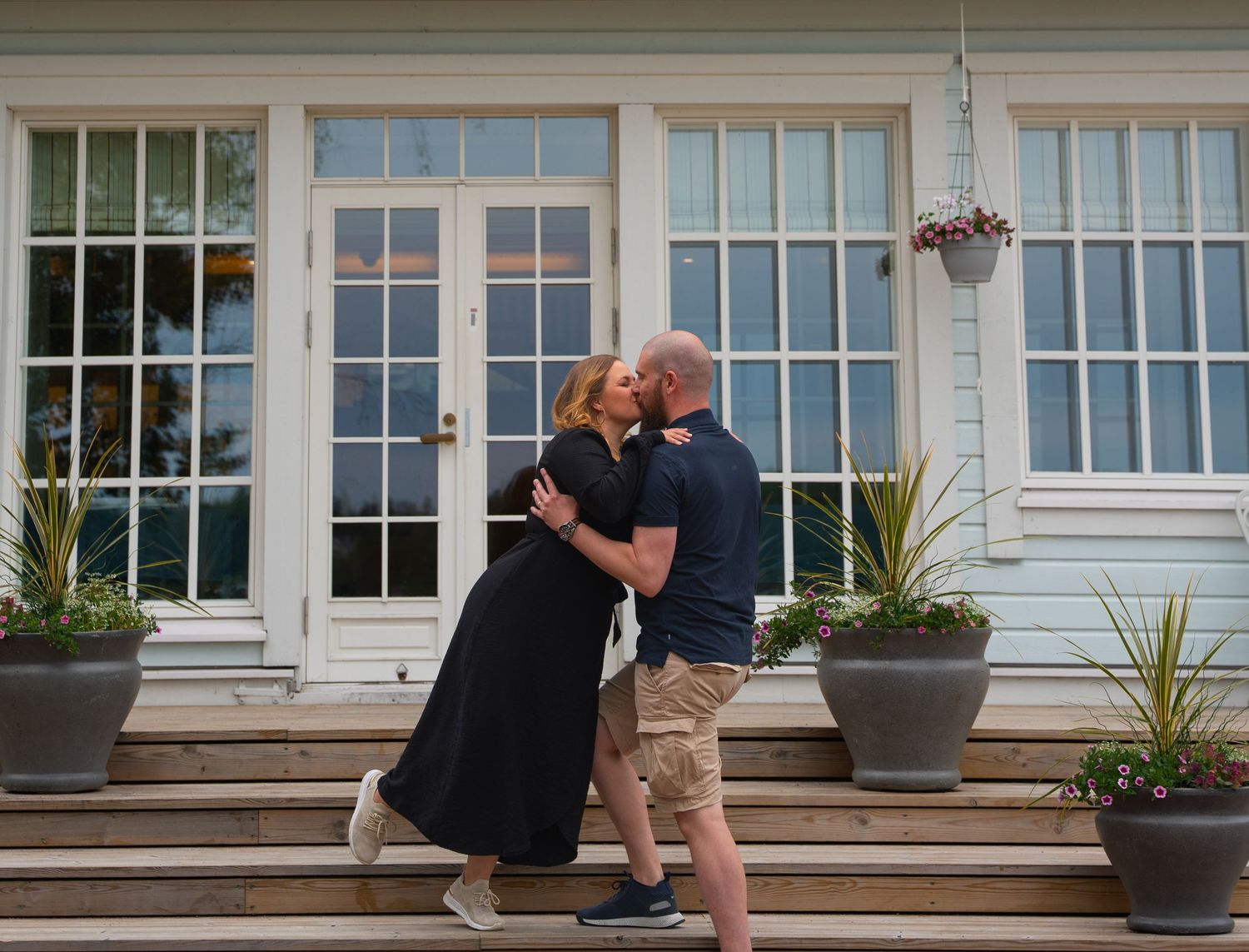 A loving couple embraces on wooden steps in front of a white house with French doors and potted plants.