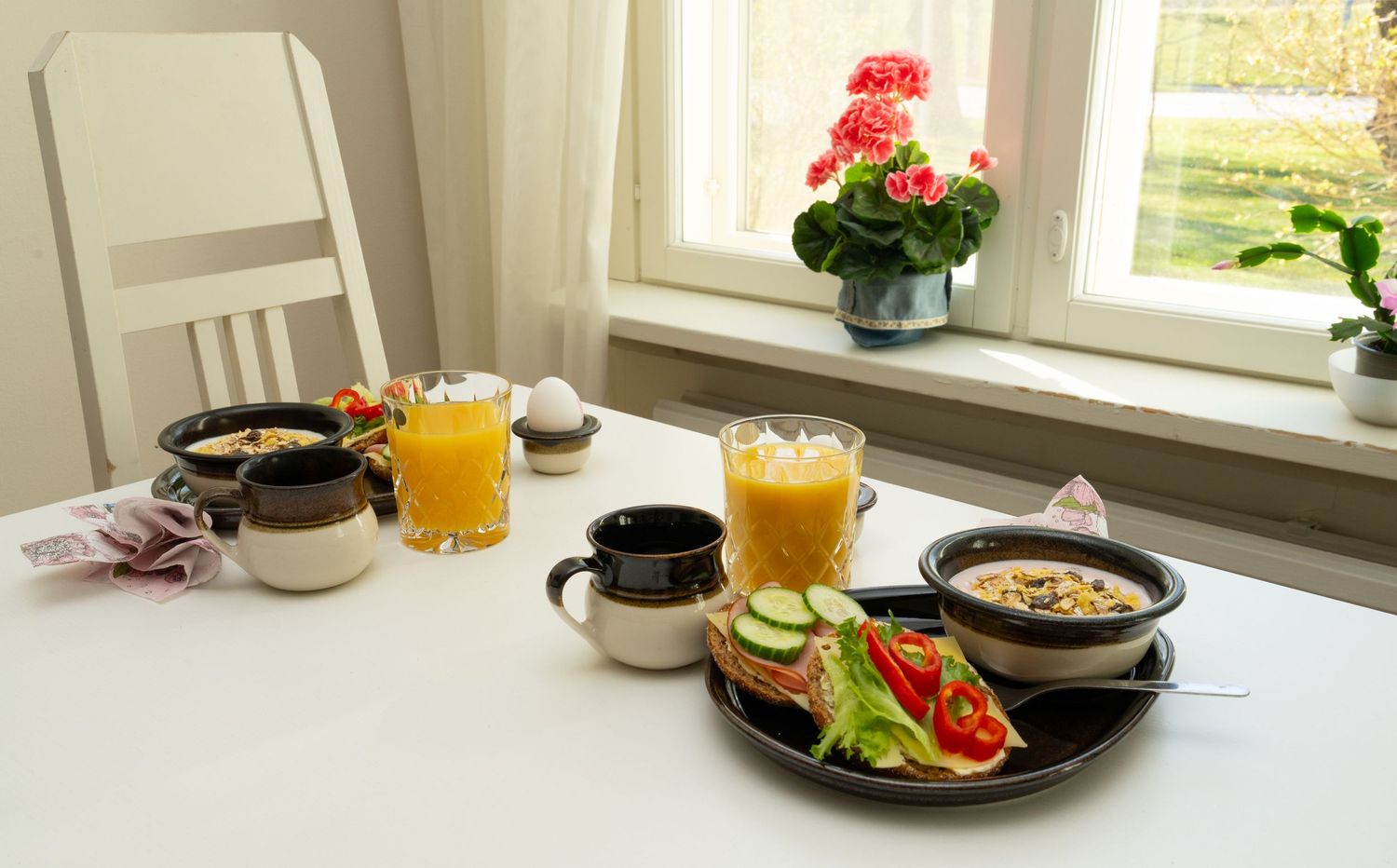 Breakfast table set with orange juice, soup bowls, fresh vegetables, and red geranium flowers by a bright window.