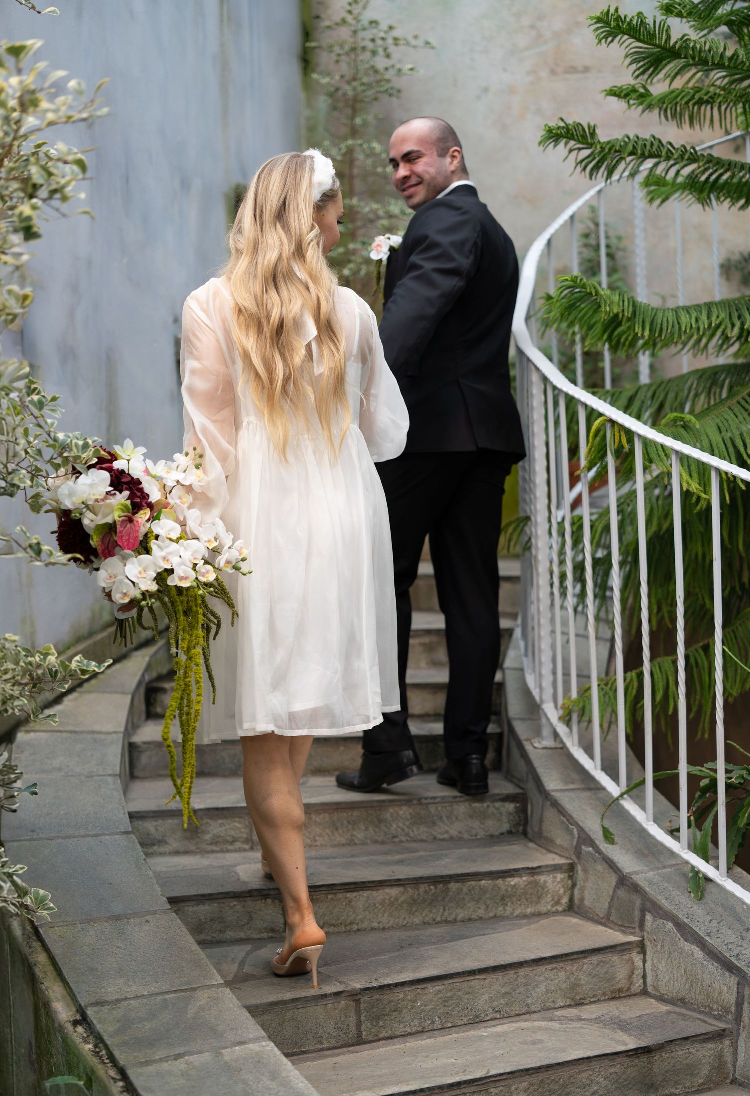 Bride in white dress holding bouquet walks up stone steps with groom in black suit at outdoor wedding venue.