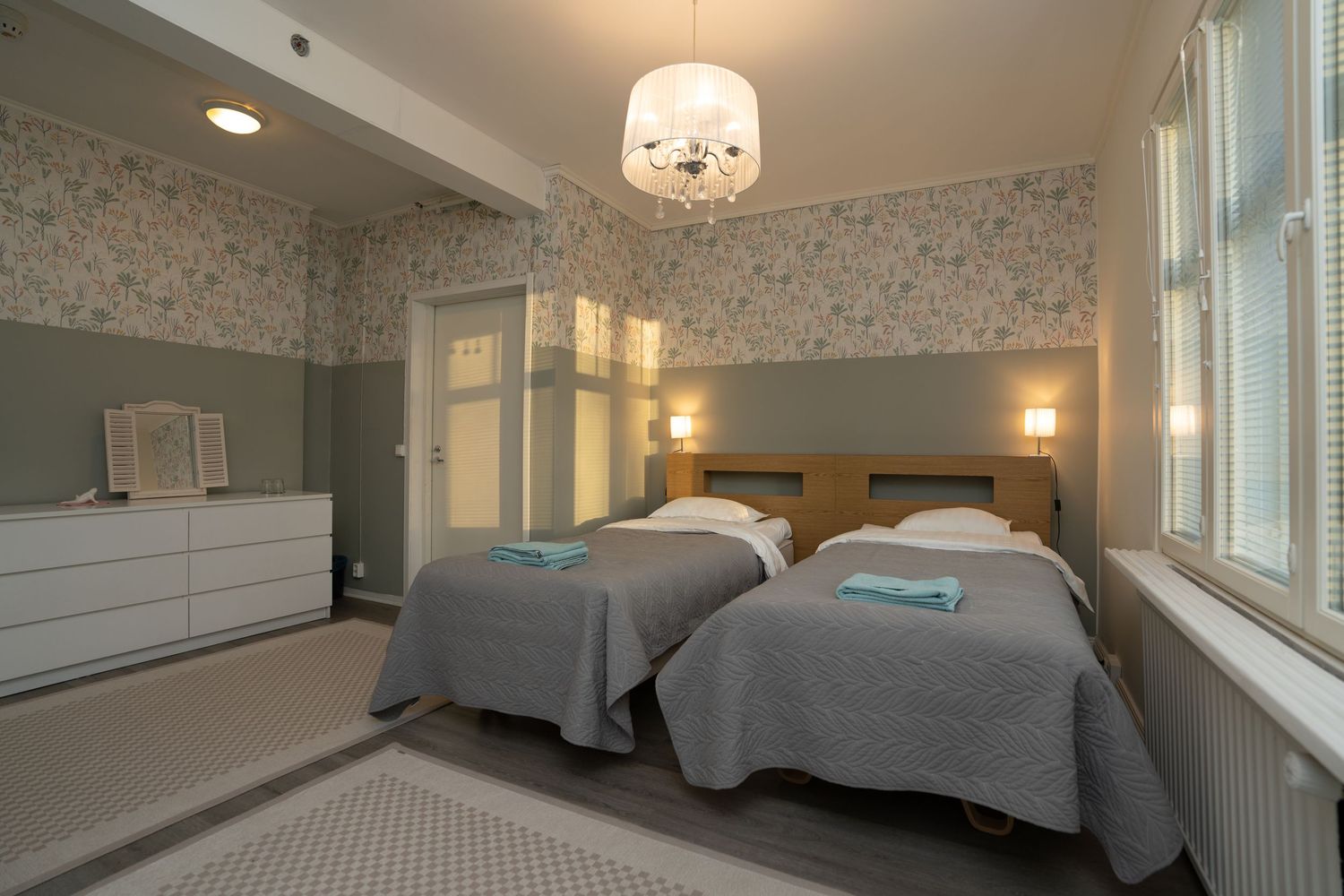 Elegant twin bedroom with floral wallpaper, chandelier, gray bedding, and teal accents in a cozy hotel room.