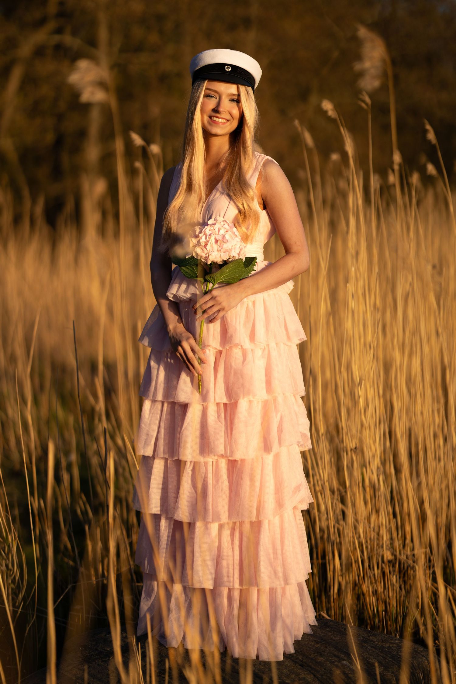 Smiling blonde woman in pink ruffled gown and sailor cap holding flowers, standing in golden tall grass at sunset.