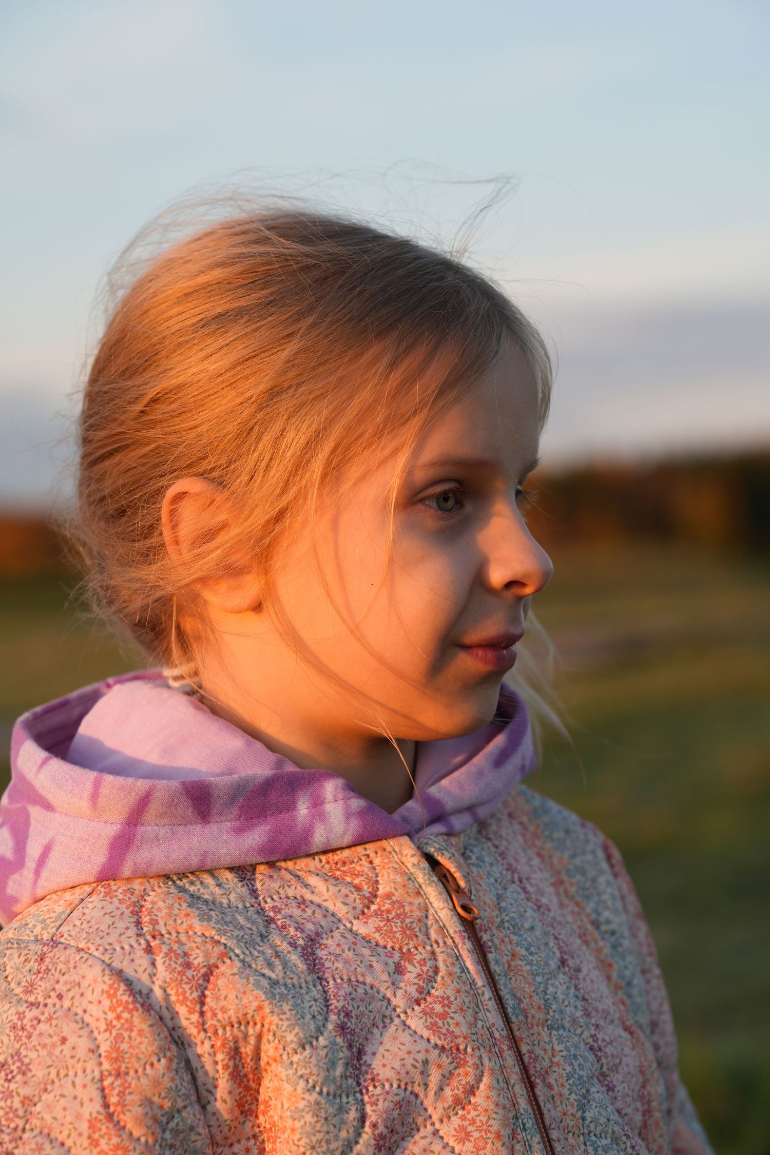 Young child in pink jacket gazes to the side during golden hour in an outdoor setting.