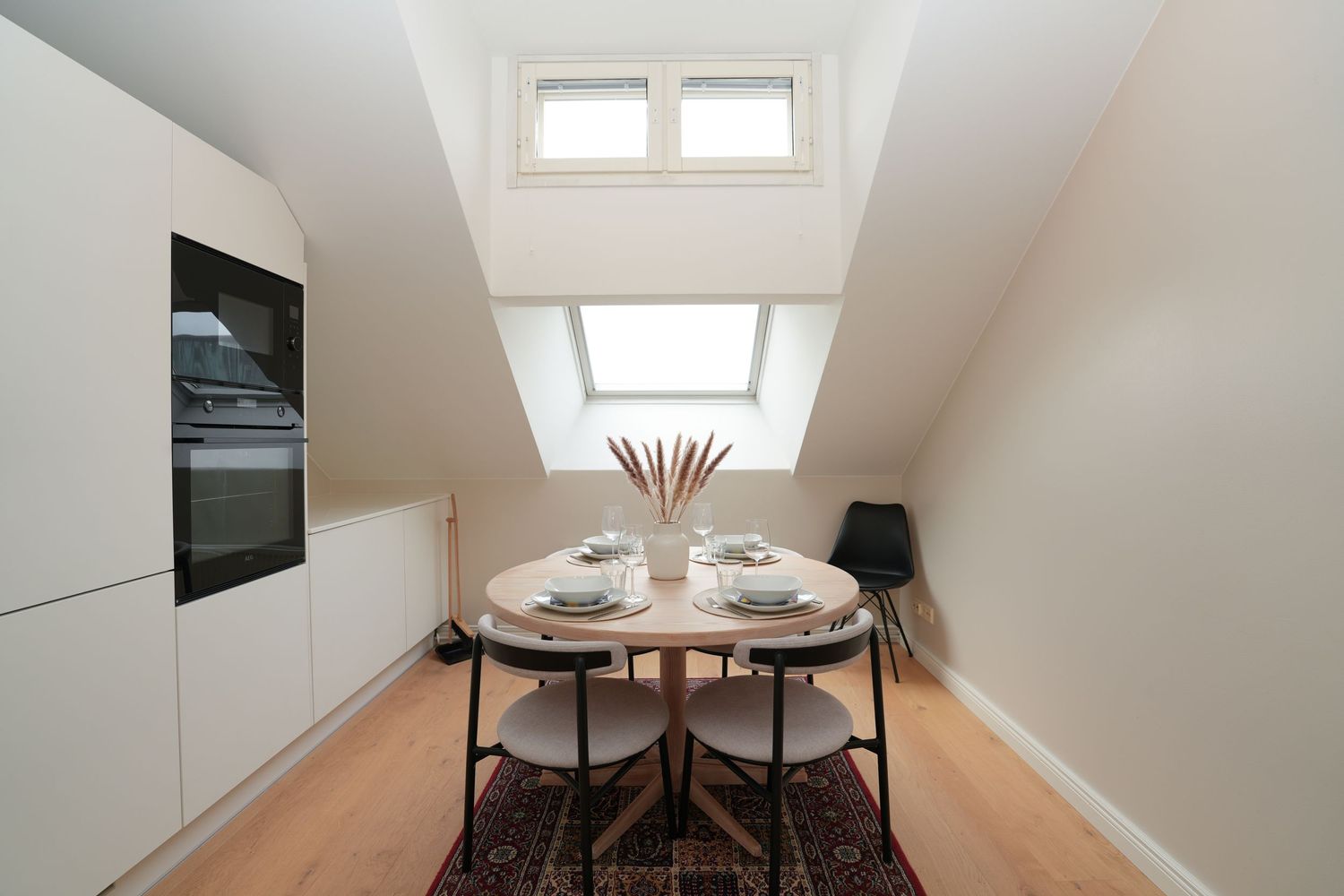 Compact attic dining area with round table, black chairs, and skylight window in modern white kitchen.