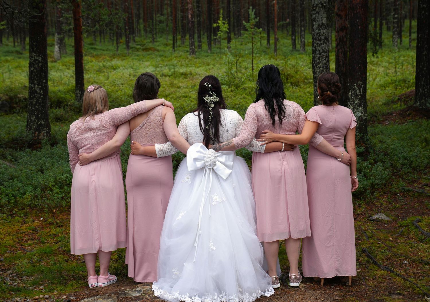 Wedding party in pink dresses and white gown photographed from behind in forest setting.