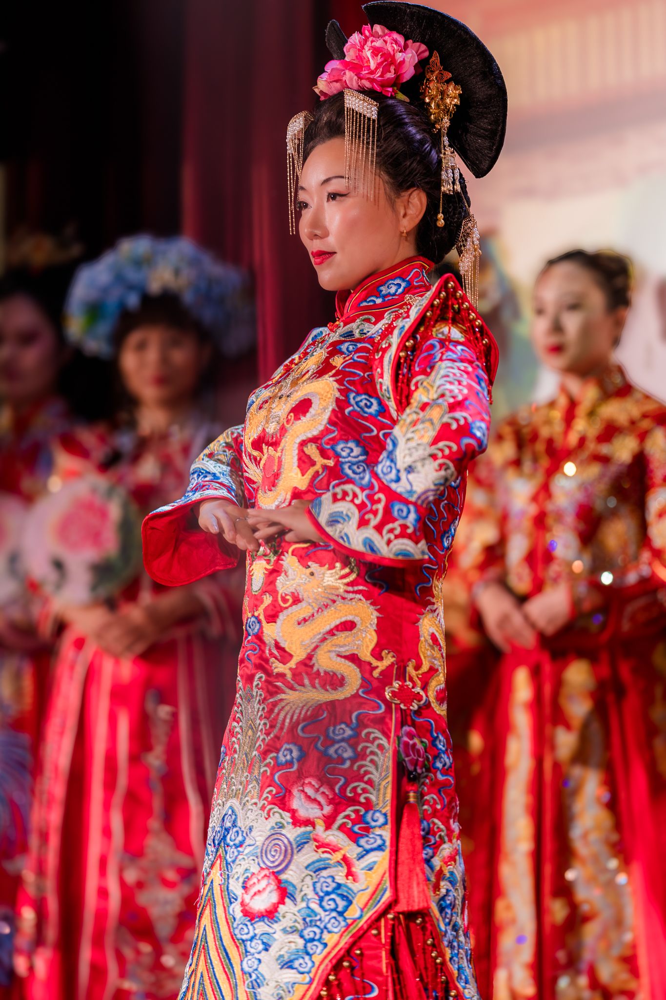 Model wearing ornate traditional Chinese qipao dress with floral headpiece on stage.