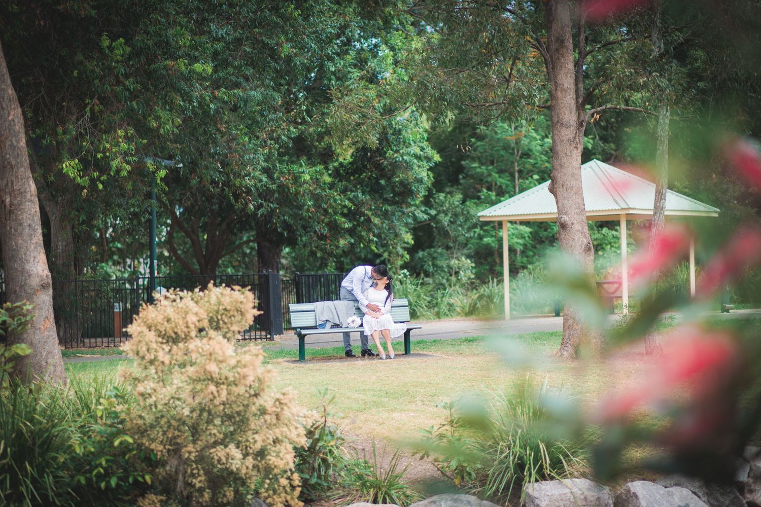 A peaceful garden scene with a bench under umbrellas surrounded by lush greenery and blooming flowers.