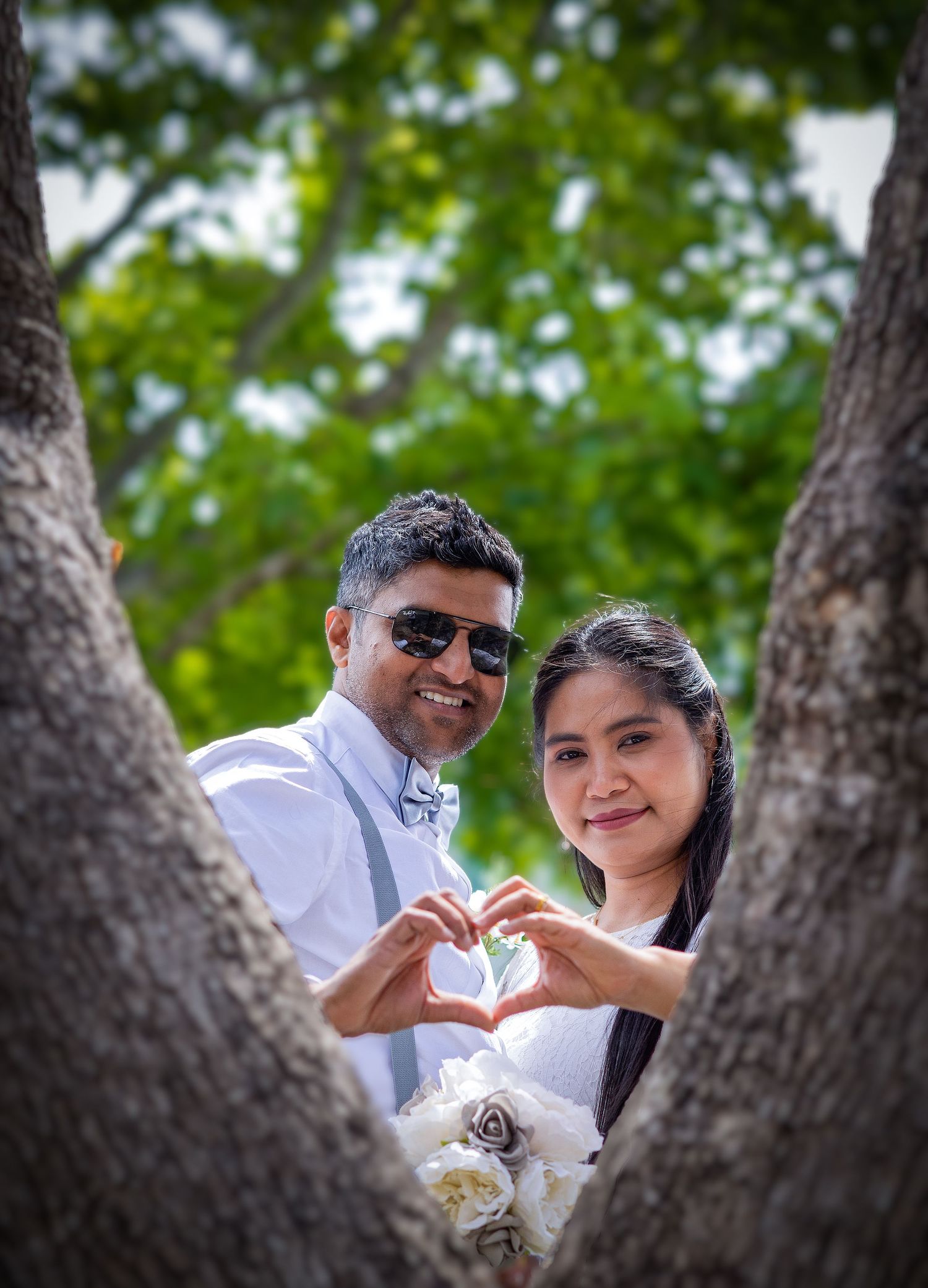 A couple poses framed between tree trunks making a heart shape with their hands in an outdoor setting.