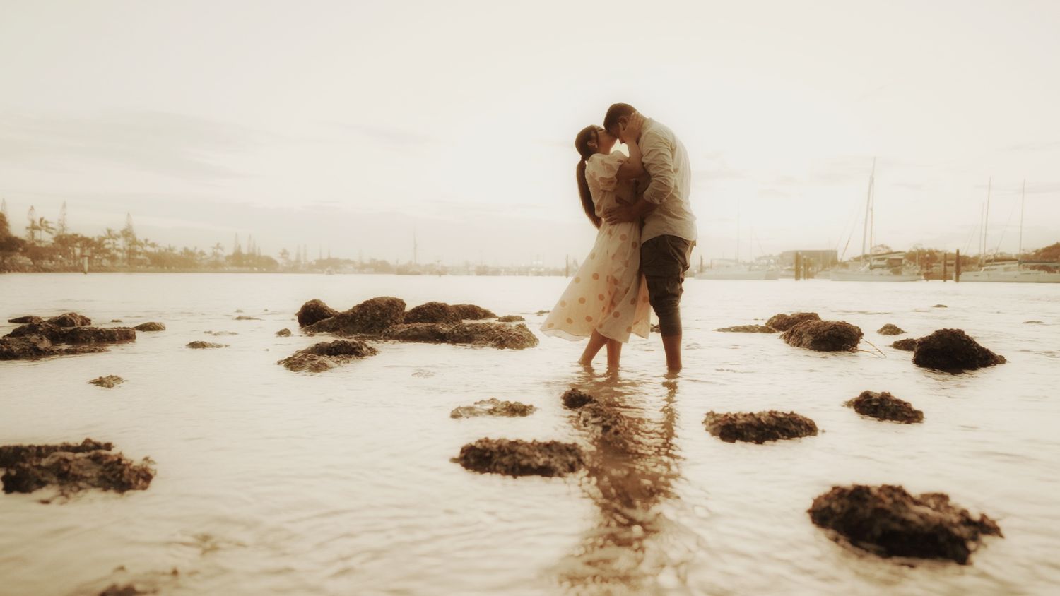 Sepia-toned romantic scene of couple standing on rocky beach during sunset.