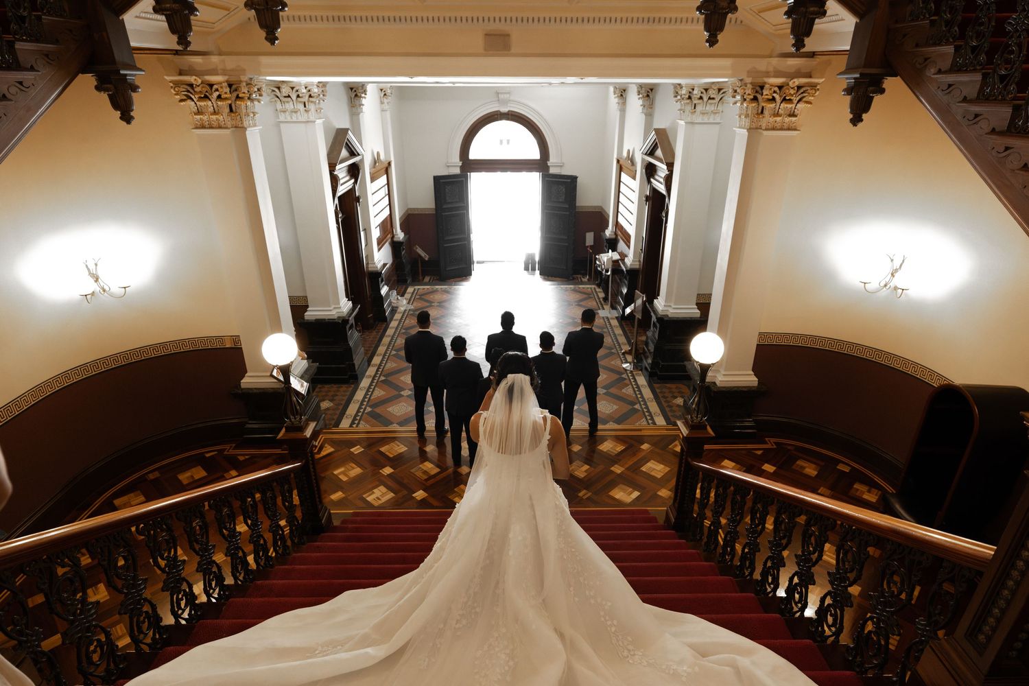 Elegant wedding scene photographed from above showing a long flowing white dress train on grand ornate staircase with lamp-lit walls.