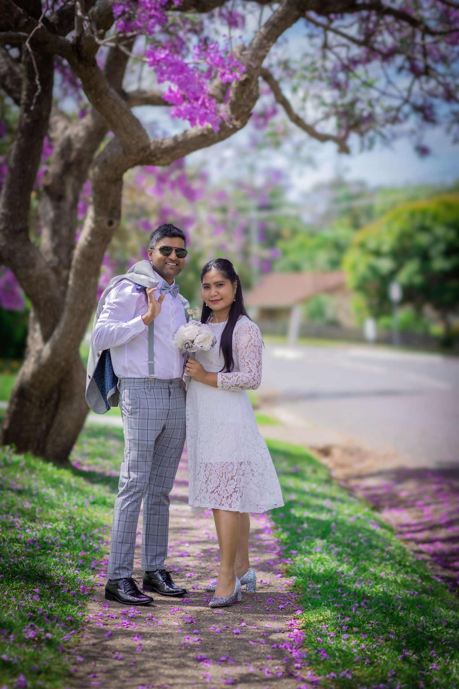 Couple posing under blooming purple jacaranda tree on suburban street.