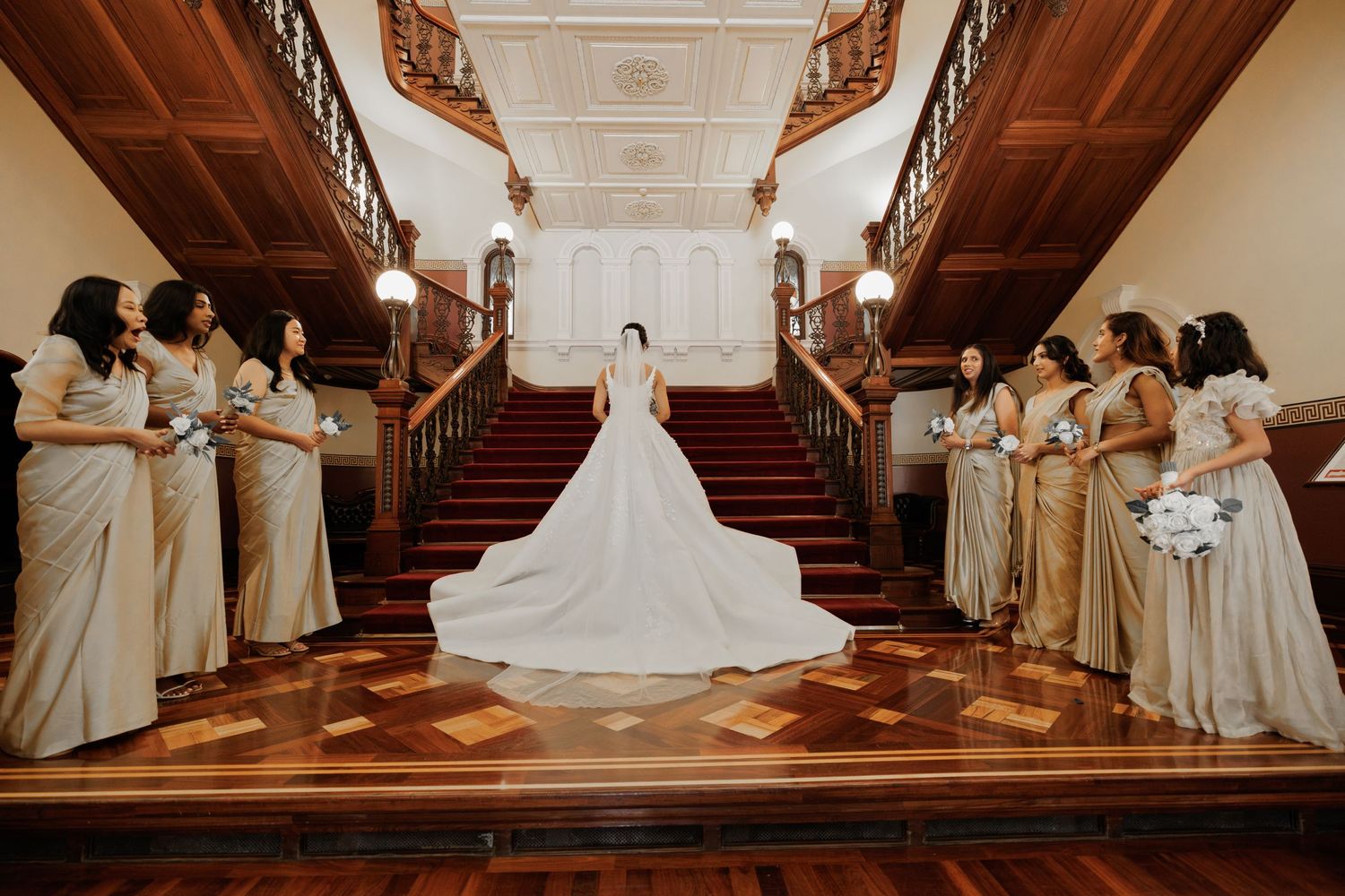A bride in a white wedding gown stands at the bottom of a grand staircase flanked by bridesmaids in champagne dresses.