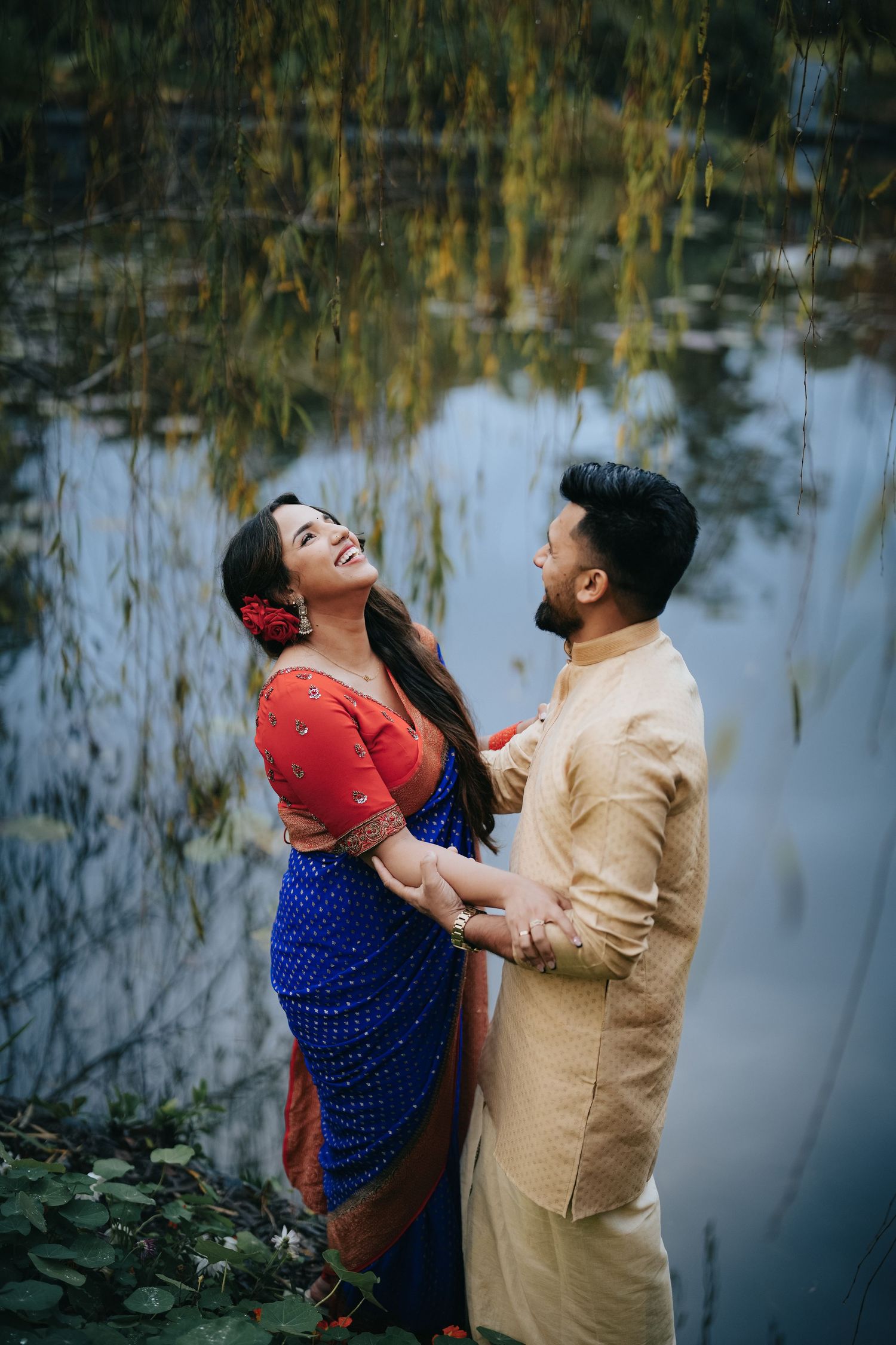 Couple in traditional Indian attire pose romantically by reflective pond with willow trees.
