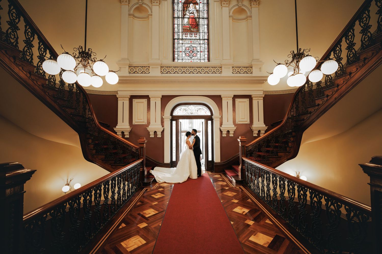 Elegant grand staircase with ornate railings and chandelier lighting leading to an arched entrance in a historic venue.