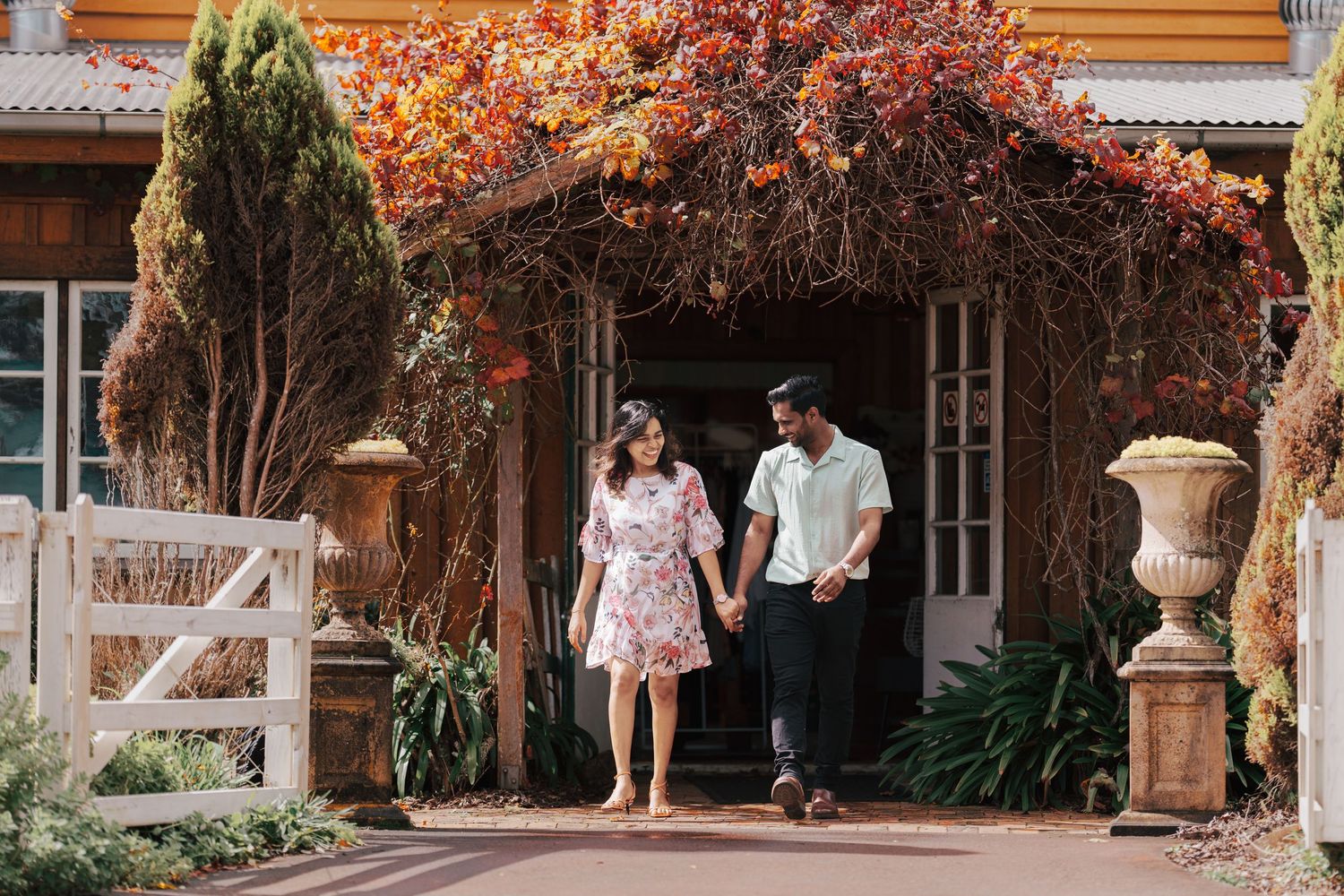 A couple stands beneath a romantic archway covered in vibrant autumn foliage at a rustic cottage entrance.