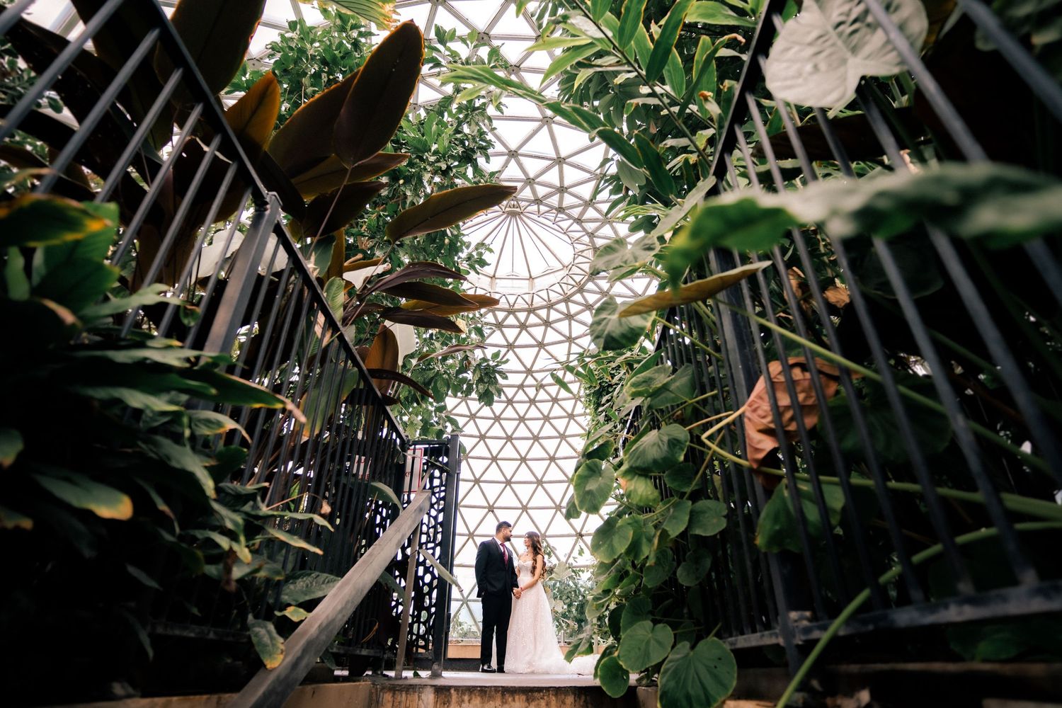 Couple walking through geodesic dome greenhouse surrounded by lush plants and architecture.