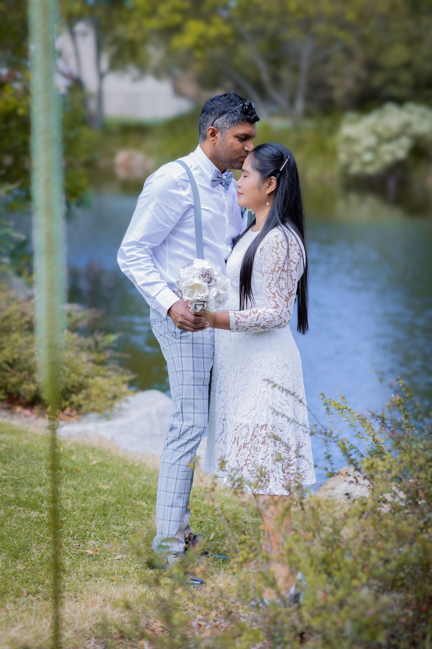 A romantic couple embraces by a lake, wearing white formal attire during an outdoor photoshoot in a garden setting.