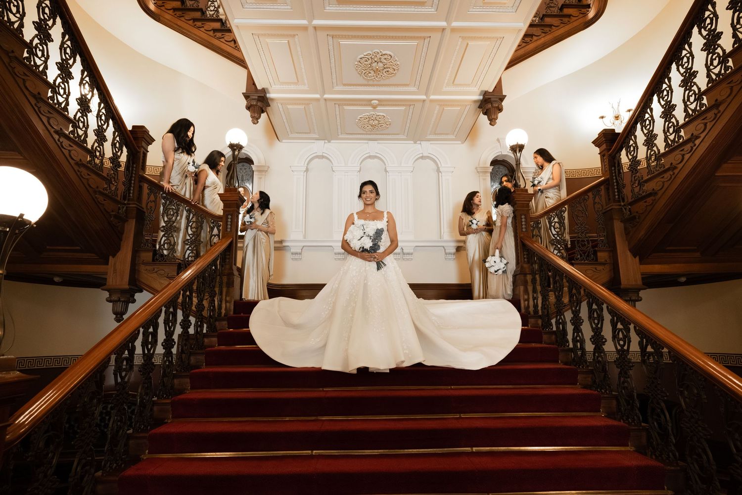 A wedding moment on an elegant wooden staircase with red carpet and ornate white walls in a historic venue.