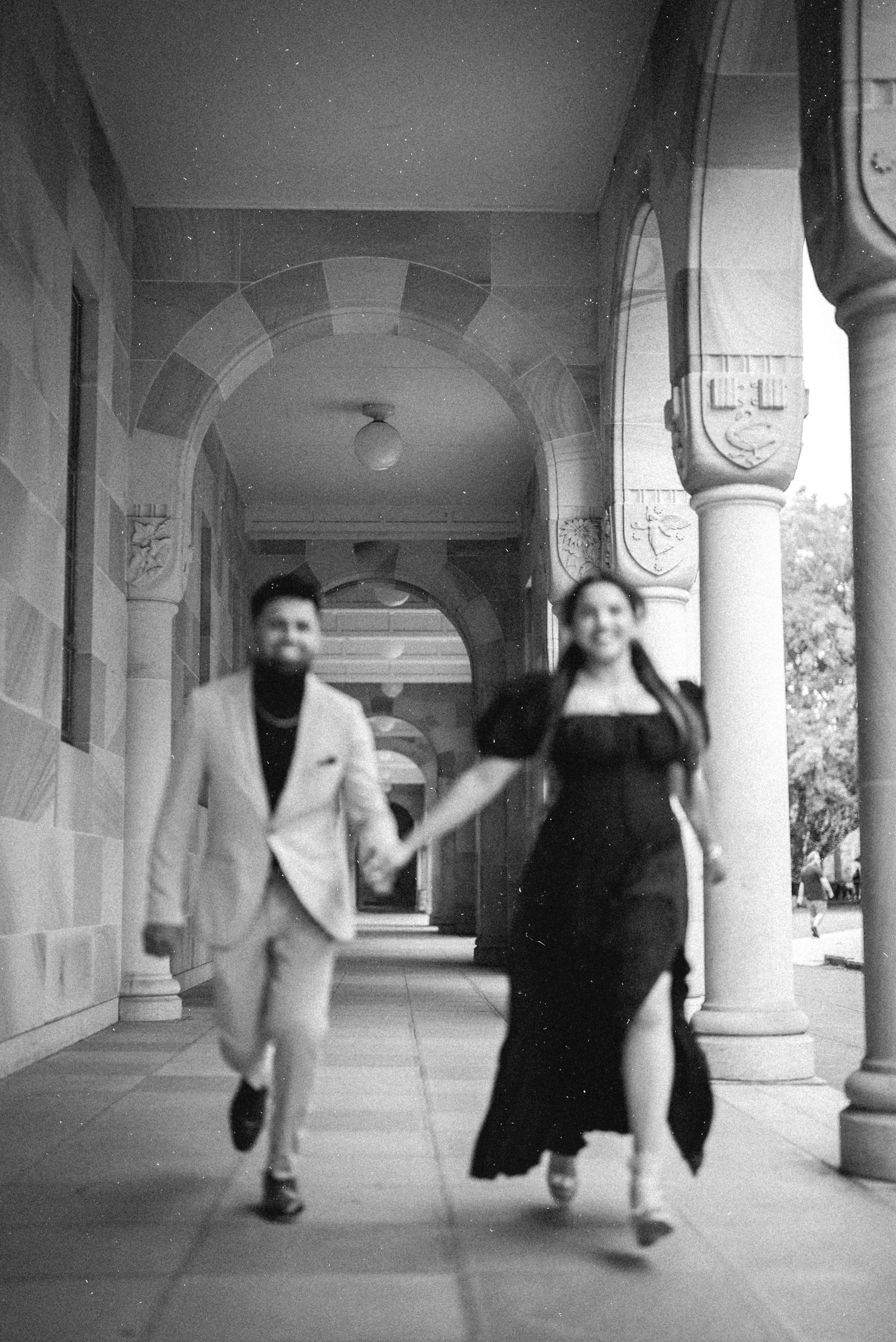 Black and white photo of couple running through elegant stone archway corridor.