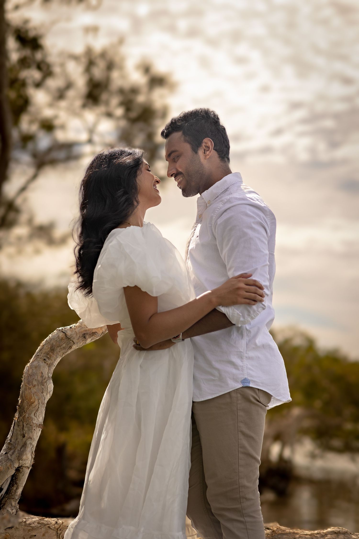 Romantic couple in white clothing embrace near water during golden hour.