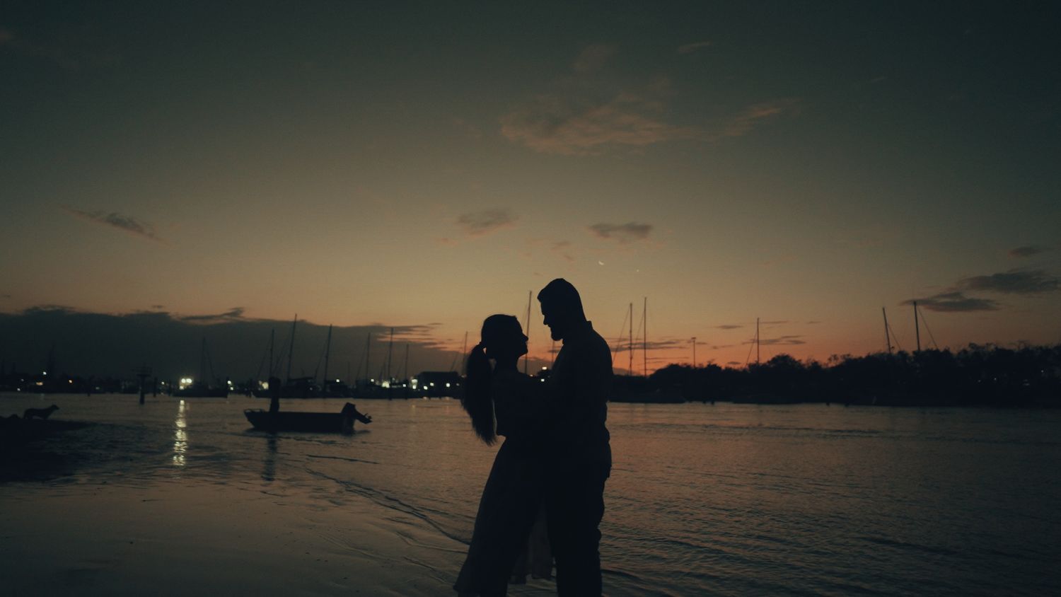 Silhouette of couple embracing against sunset over harbor with boats in background.