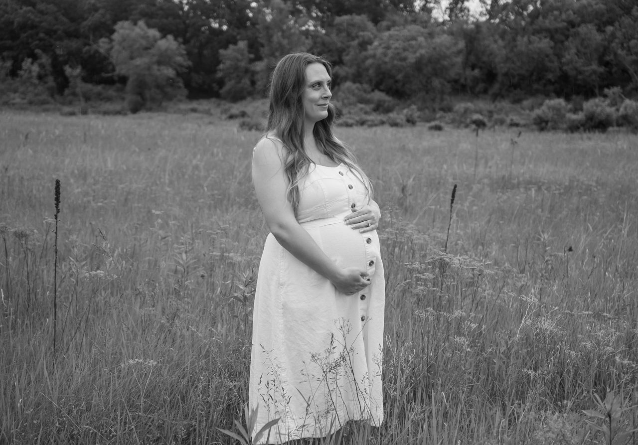 Pregnant woman in white lace dress stands contemplatively in wildflower meadow during rustic outdoor photo session.