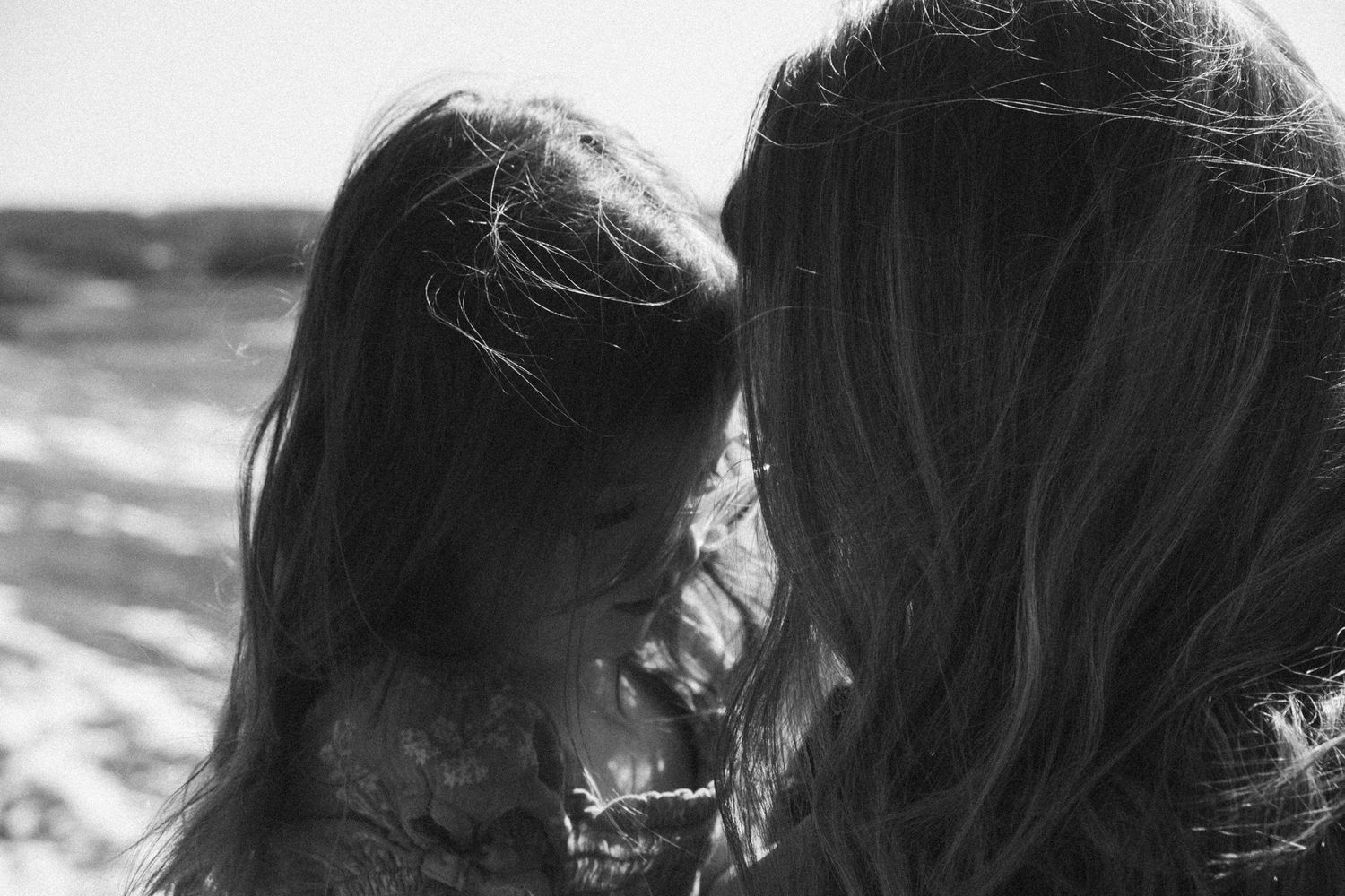 A close up black and white photo of two people facing each other with wind blowing through their hair near the ocean.