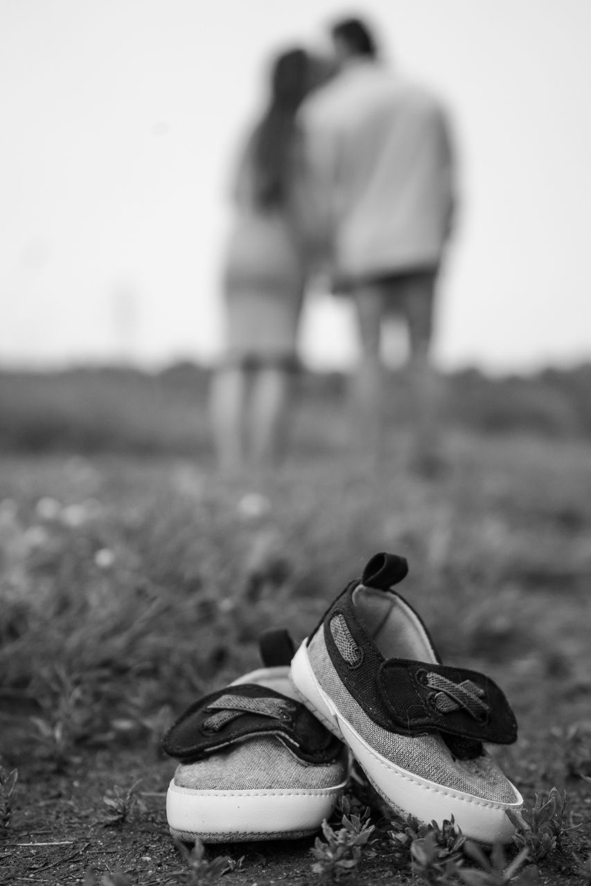 Small baby shoes in foreground with blurred silhouette of couple standing in distance, shot in black and white.