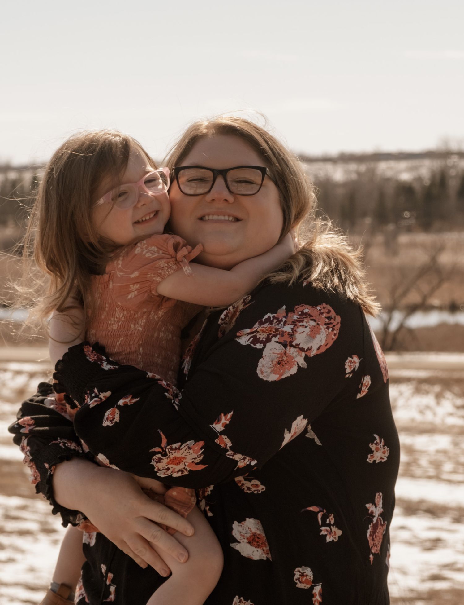 A warm winter portrait of two people embracing outdoors, with one wearing a black floral top against a snowy landscape.