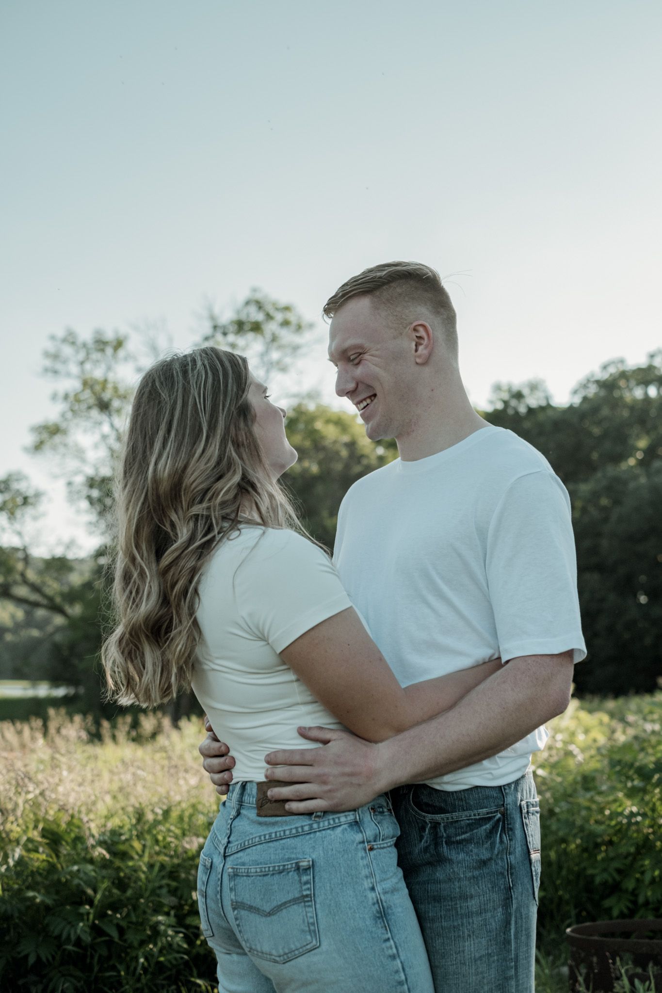 A couple wearing matching white t-shirts and jeans embrace in a sunlit field during a romantic photoshoot.
