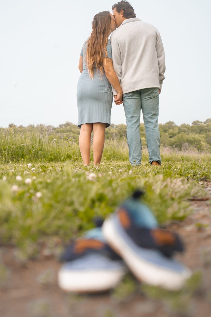Couple standing in flower field with baby shoes in foreground, shot from behind.