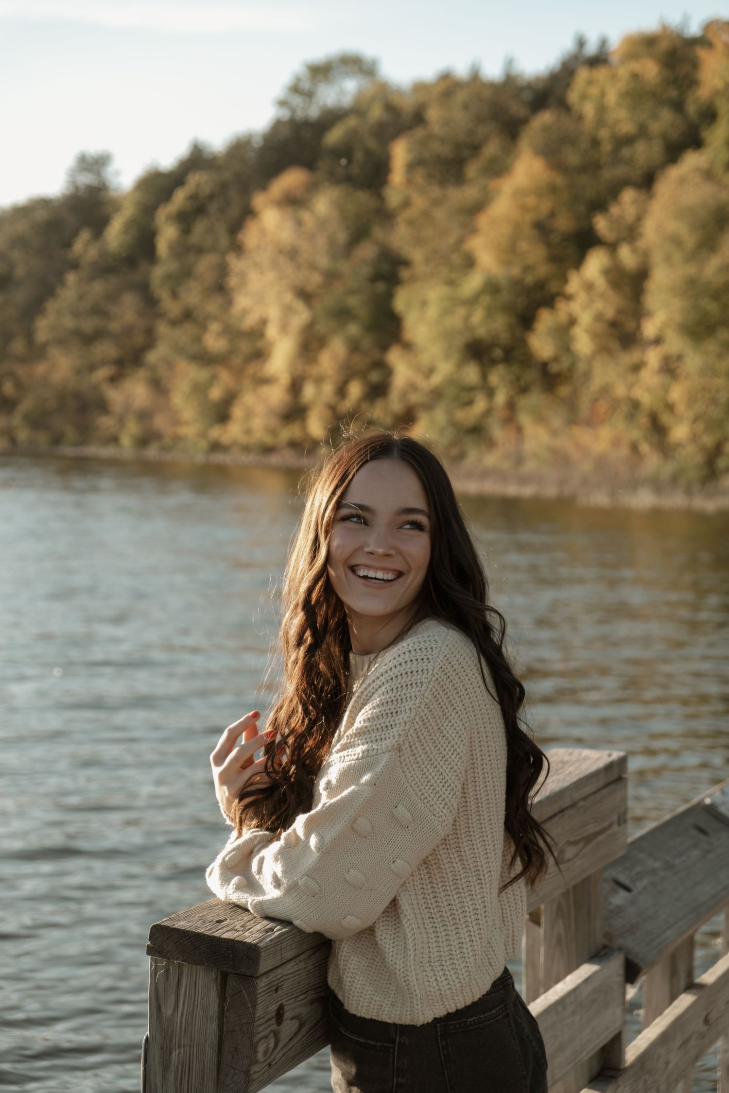 Someone with long wavy brown hair wearing a cream sweater smiles while leaning on a wooden railing near a lake.