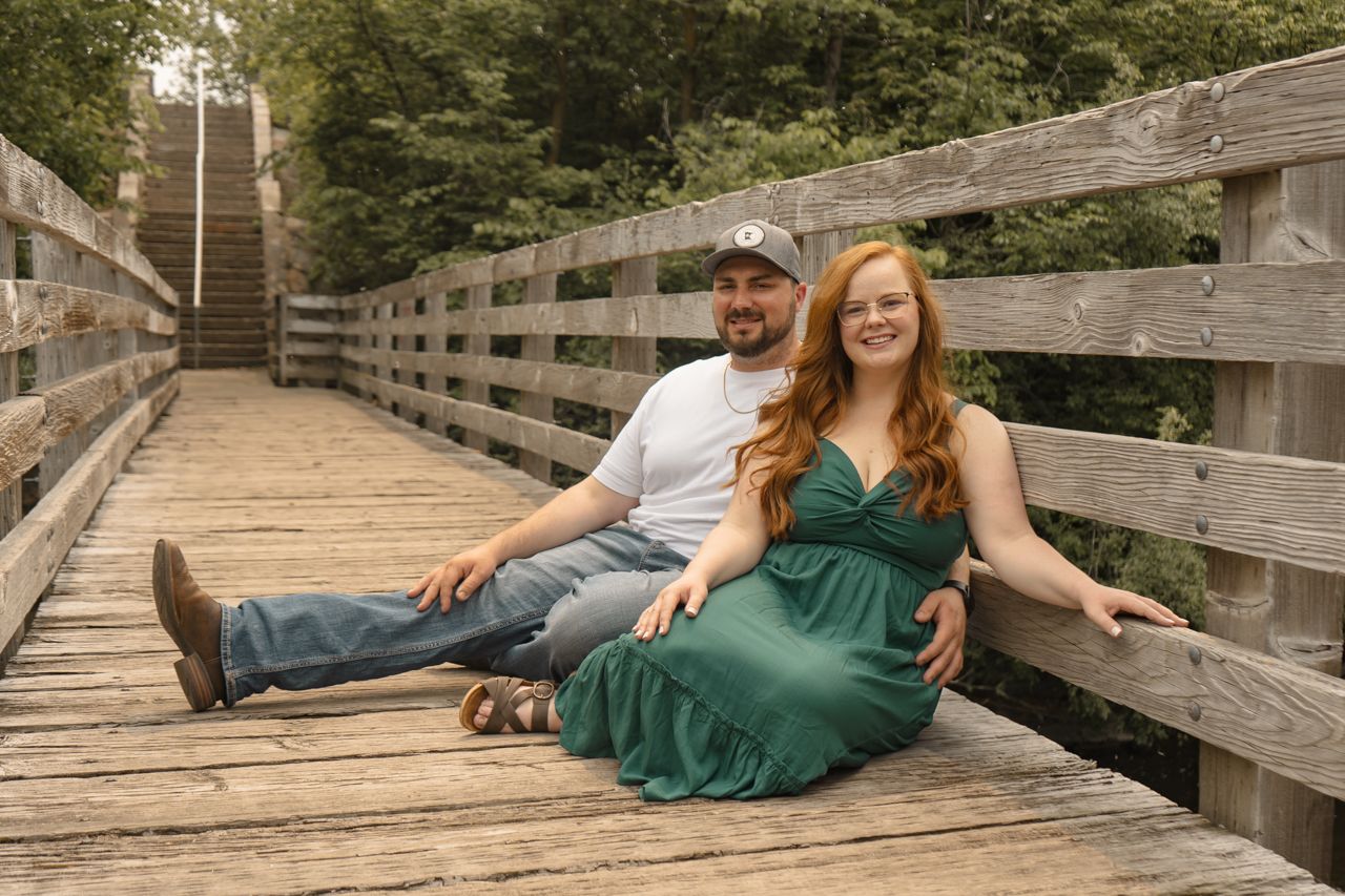 Two people relax together on a wooden bridge walkway surrounded by lush green trees on a sunny day.