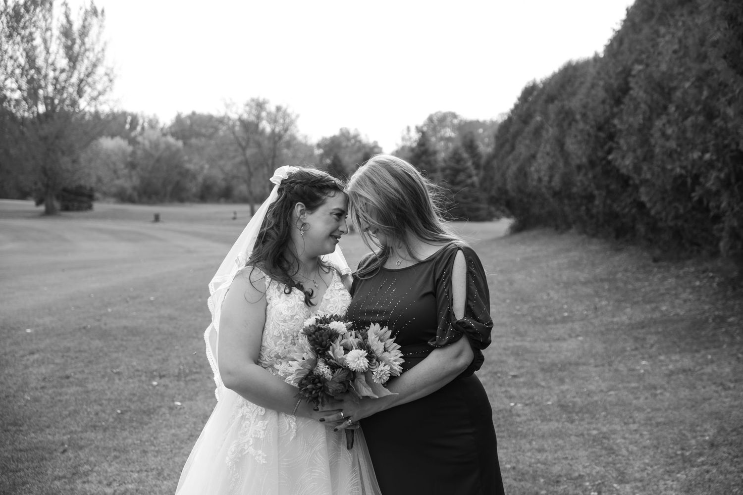 Two people share a tender moment while holding a floral bouquet in a black and white outdoor photograph.